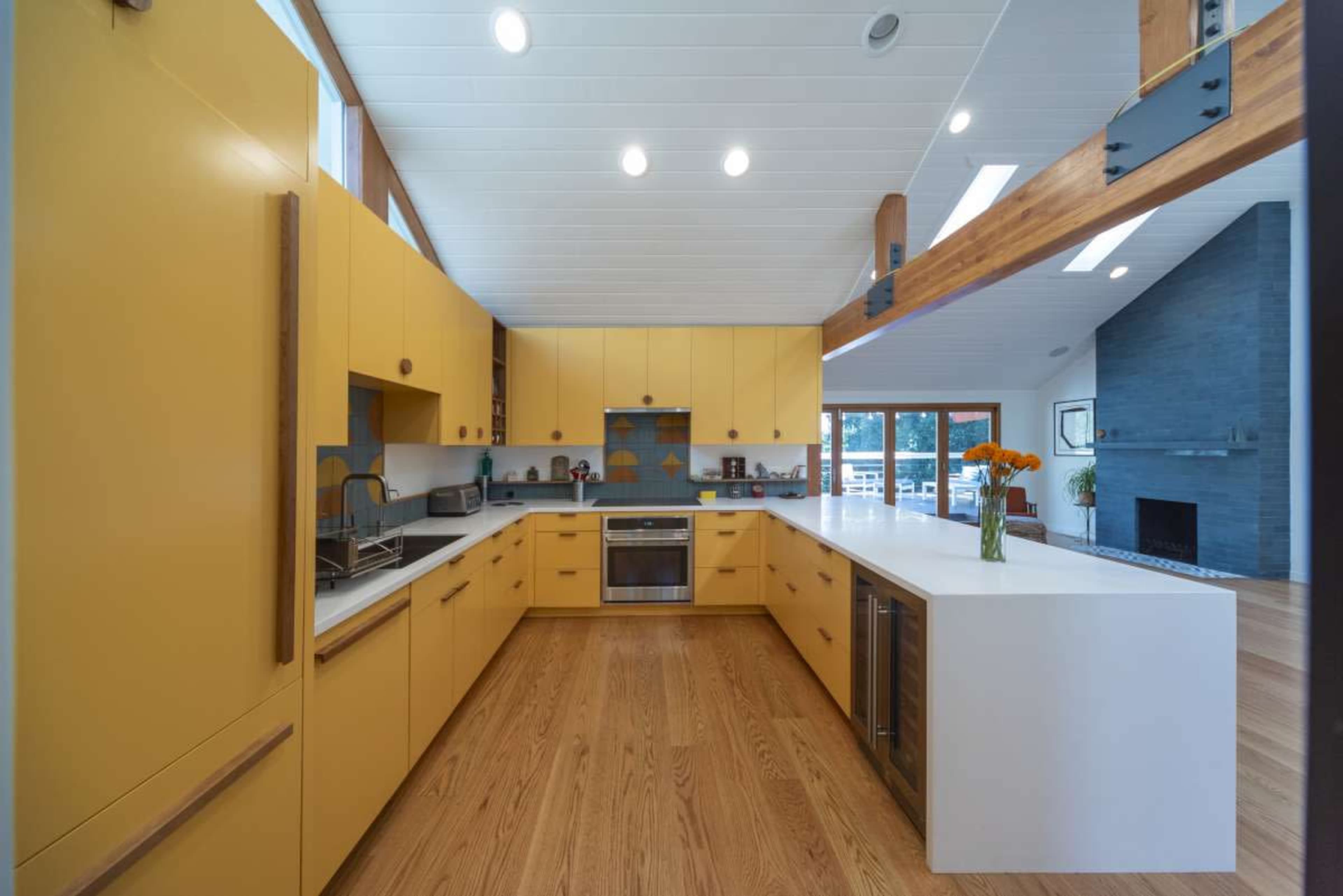 The image shows a modern kitchen with yellow cabinetry, white countertops, and wooden flooring, featuring an open space layout.
