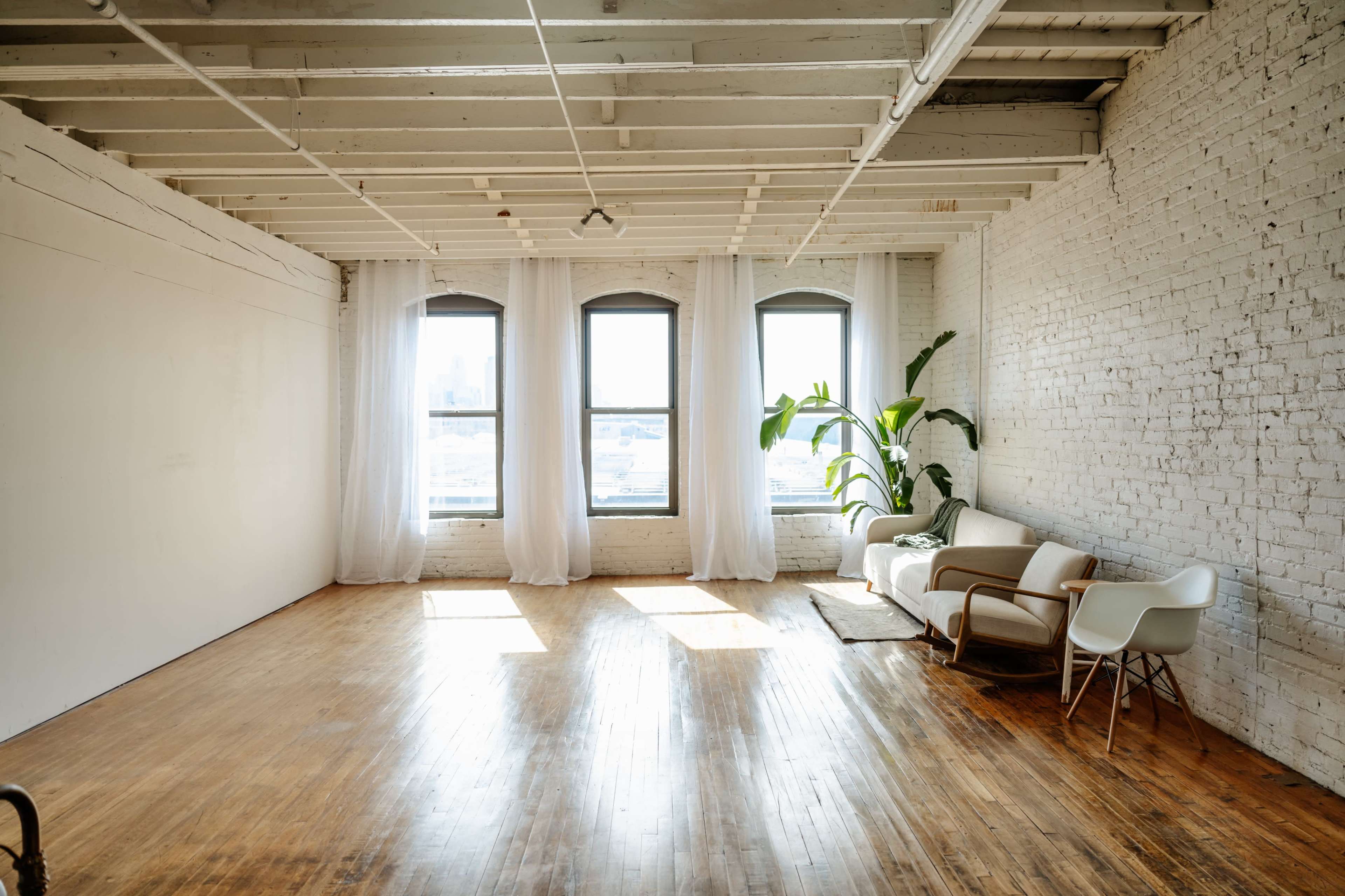 A spacious room with large windows, wooden flooring, white brick walls, and minimal furniture including a sofa and a chair.