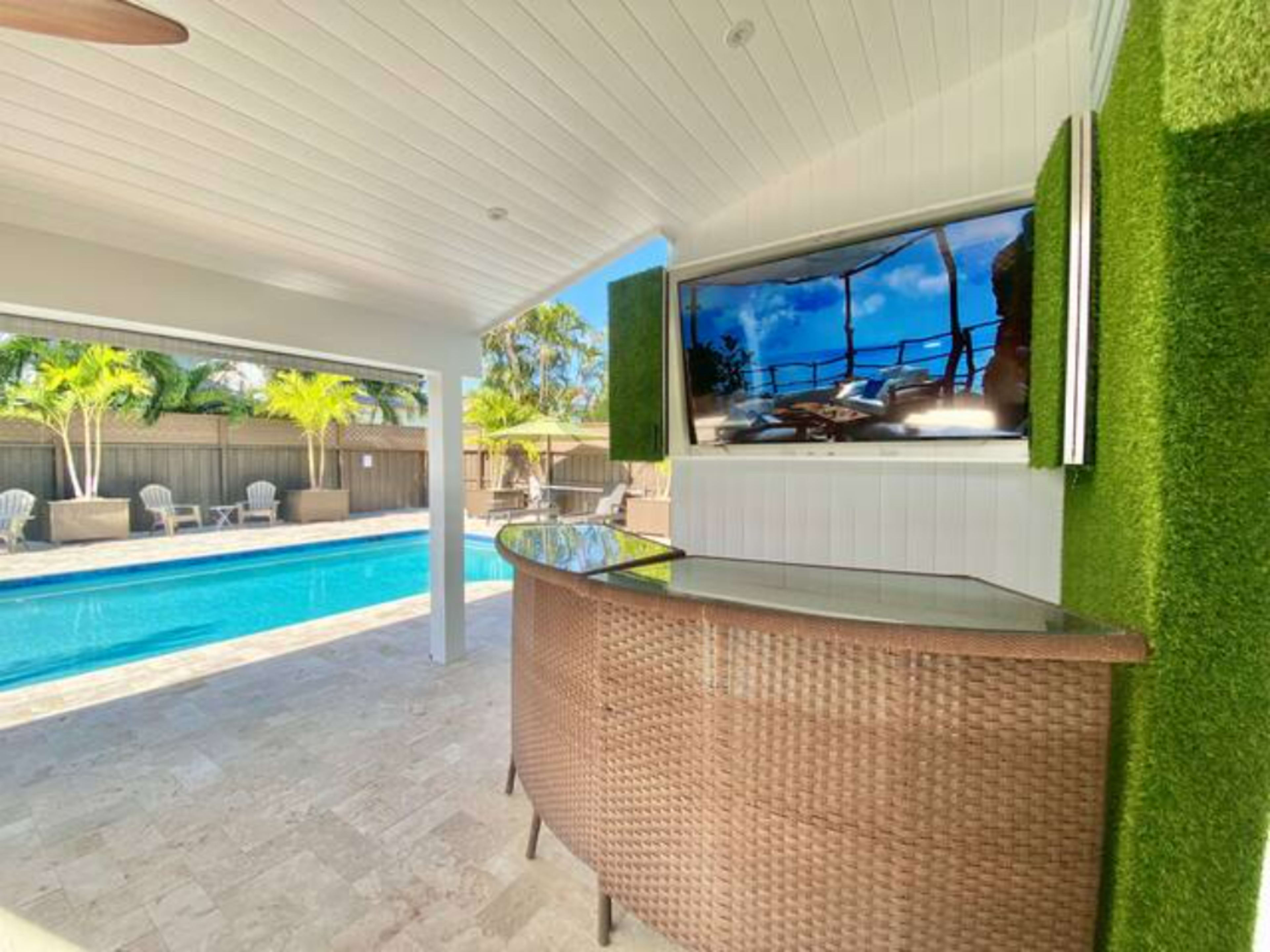 The image shows a modern outdoor bar area with a large mounted TV, overlooking a swimming pool surrounded by palm trees.