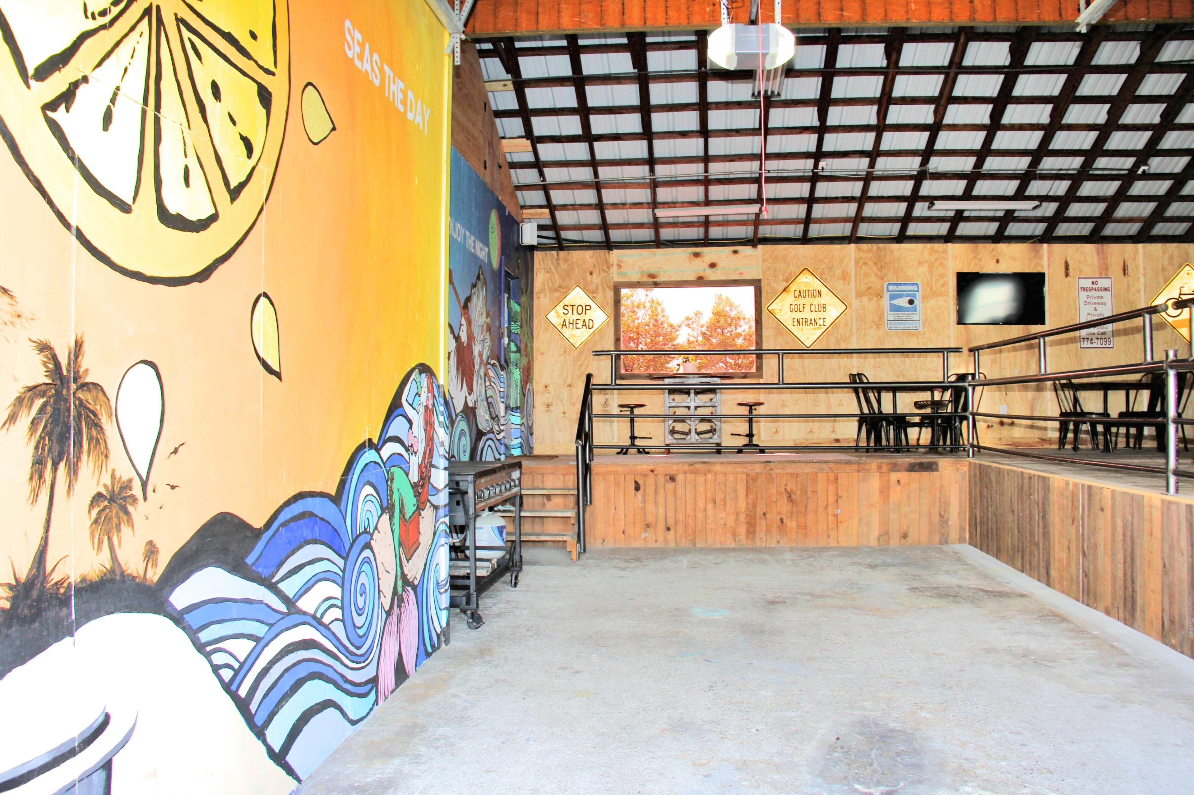 The interior of a spacious venue features colorful murals on the walls, wooden flooring, and seating arrangements with tables and chairs.