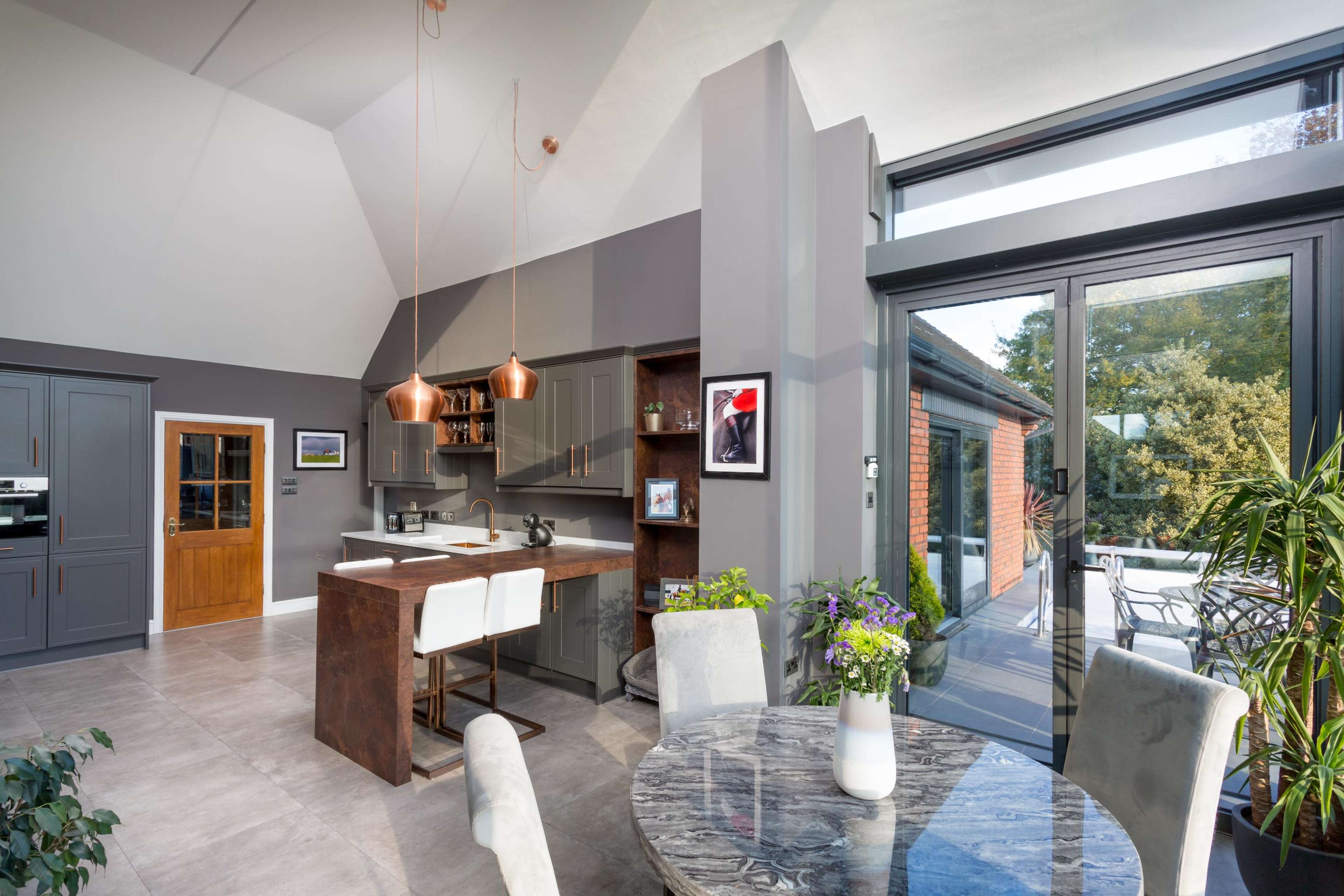 The image shows a modern kitchen and dining area featuring a dark gray color scheme, a large window, and a round table surrounded by light-colored chairs.
