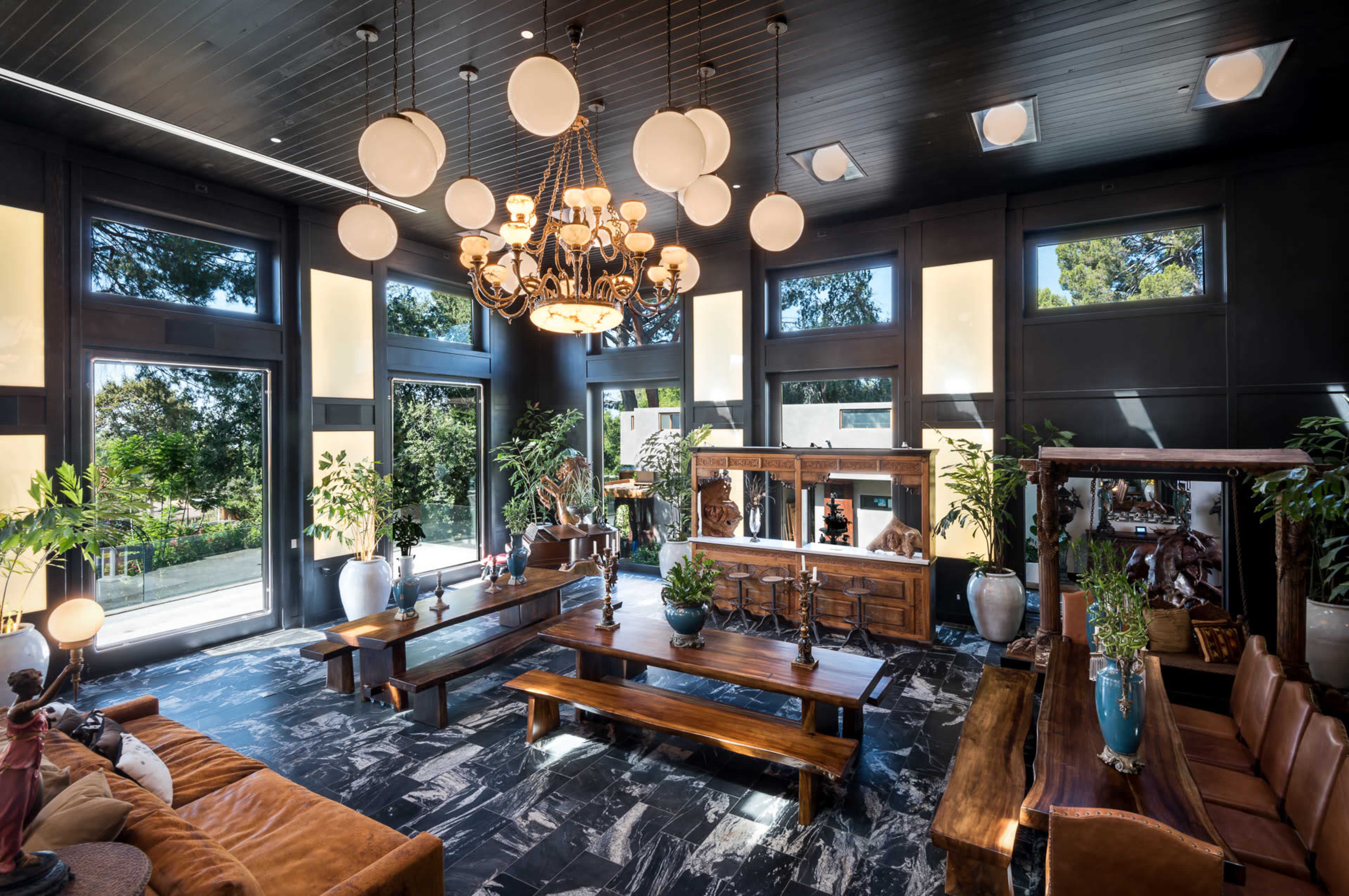 A spacious living room with large windows, a central chandelier, wooden tables, and decorative greenery.