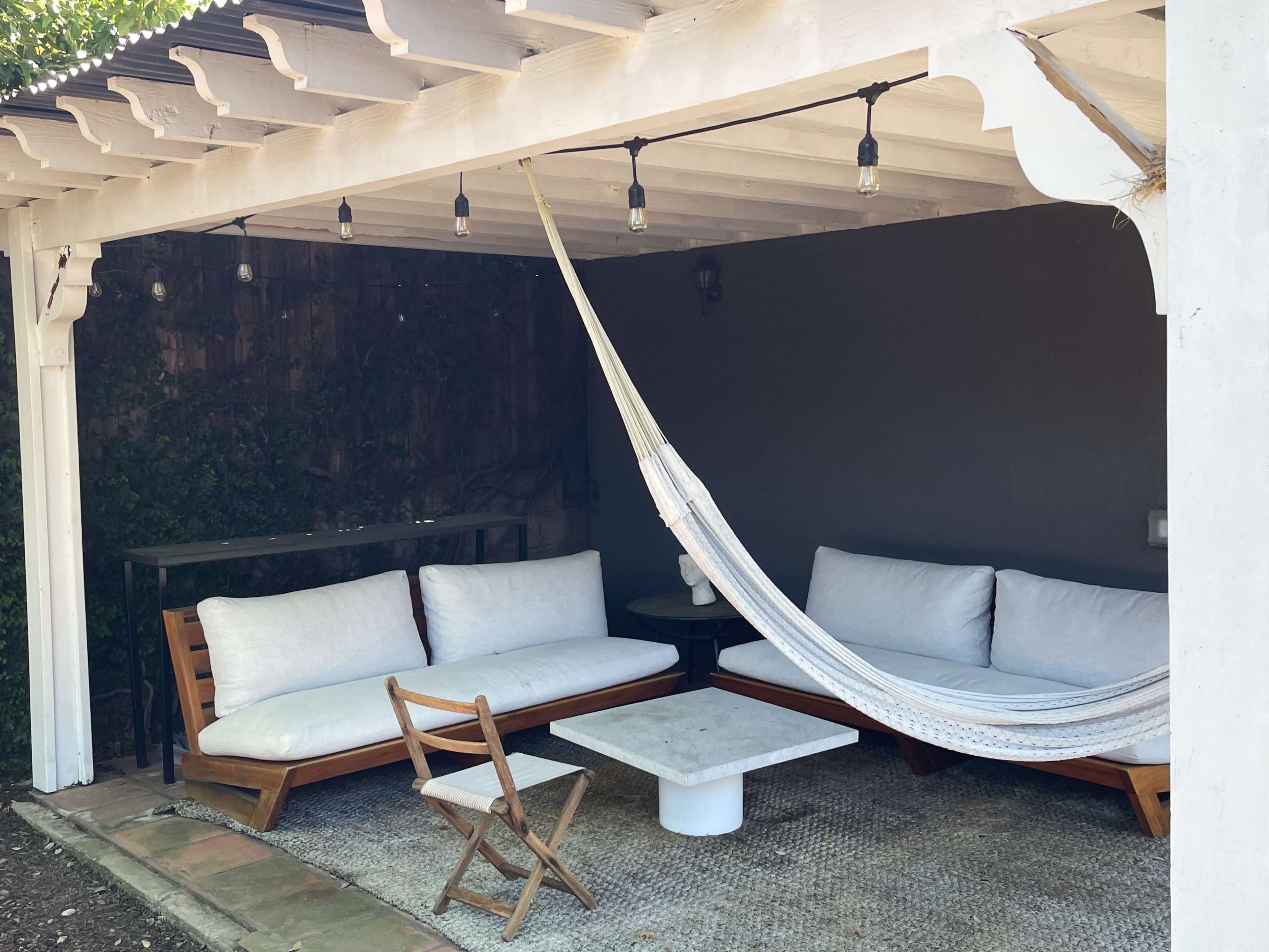 A shaded outdoor seating area features a hammock, two sofas, a small table, and a folding chair under string lights.
