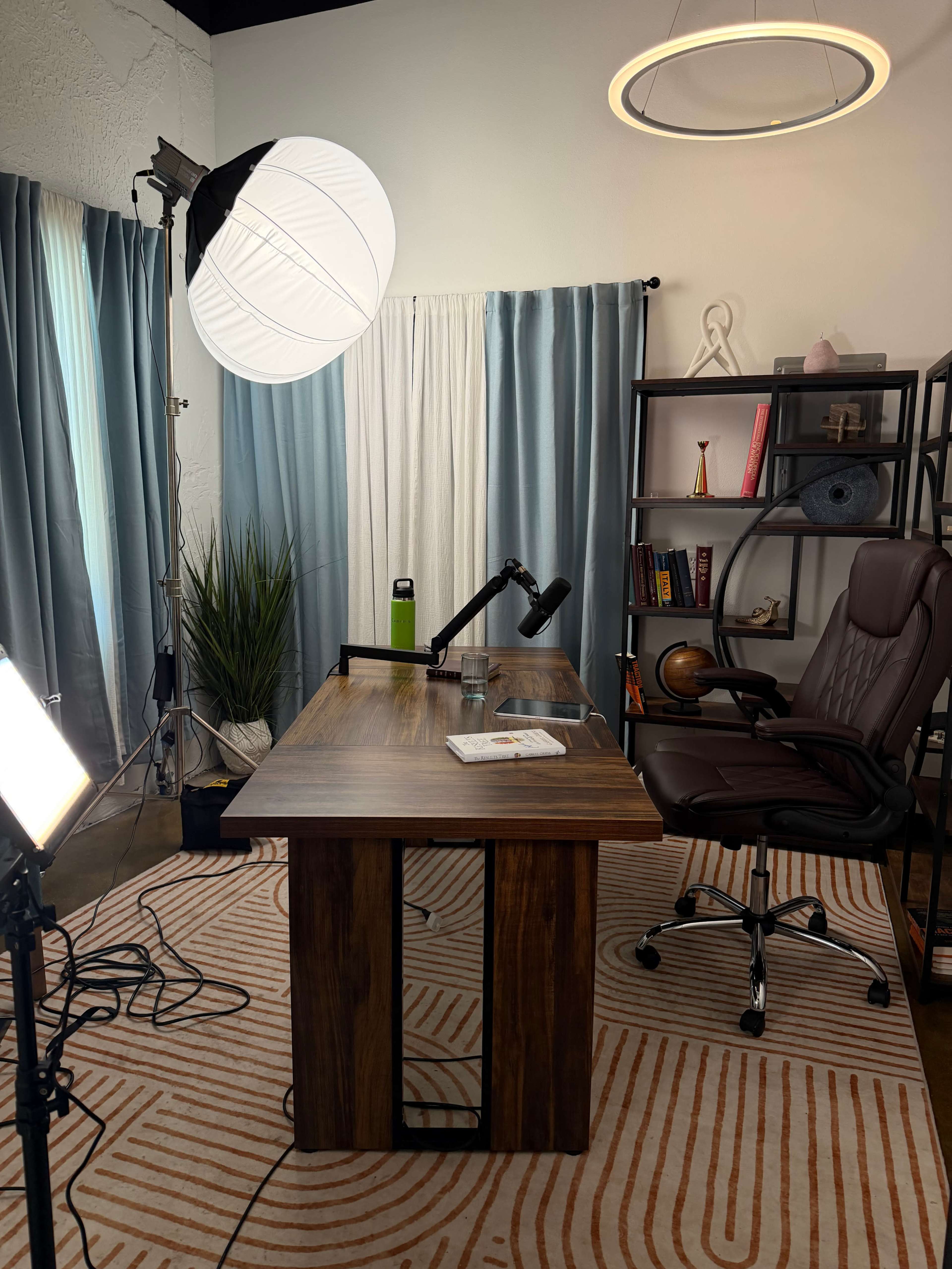 The image depicts a modern office setup featuring a wooden desk with a lamp, an ergonomic chair, and light fixtures, surrounded by curtains and decorative items on shelves.