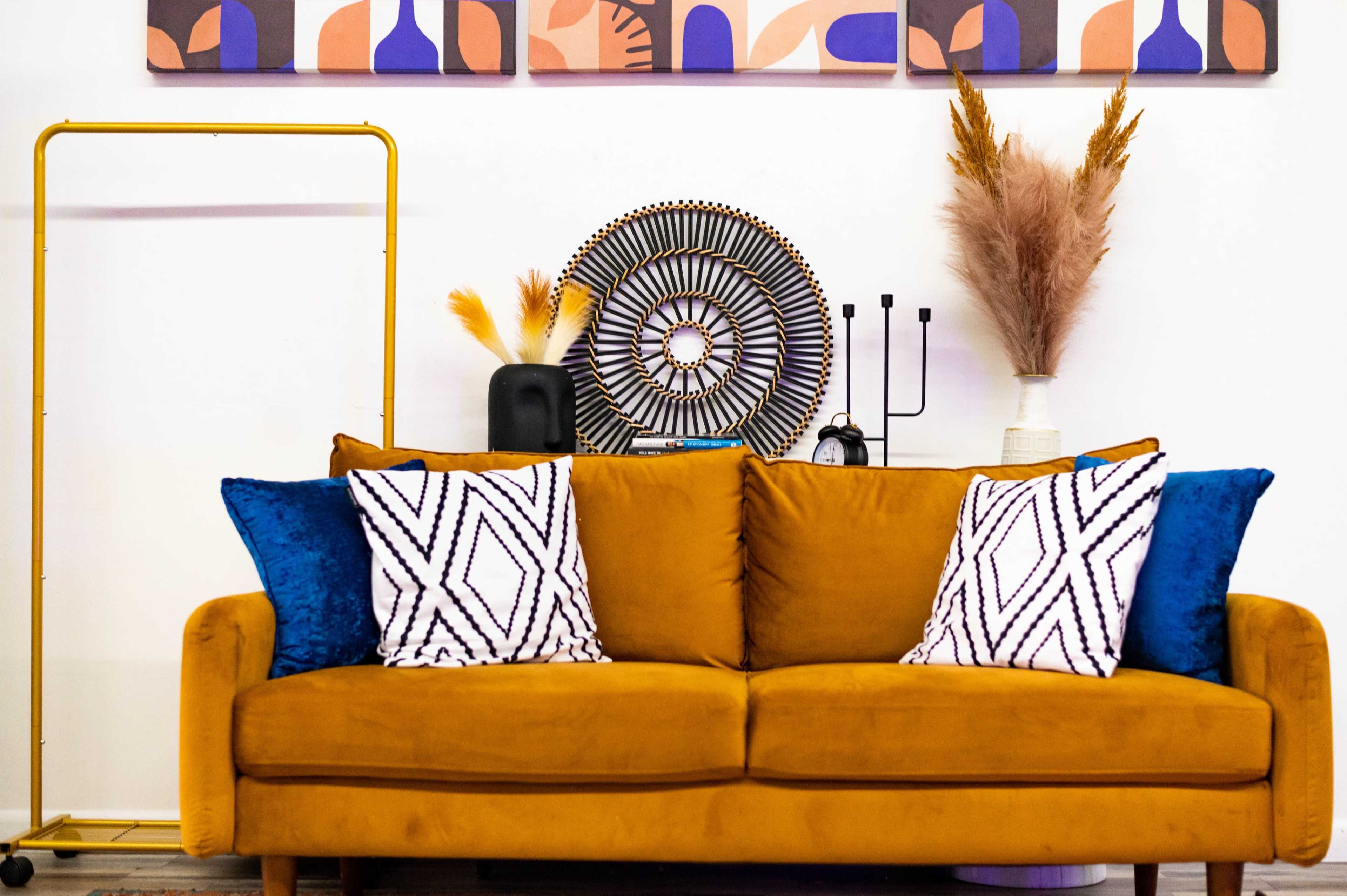 A mustard yellow sofa with patterned cushions is positioned against a wall featuring abstract art, accompanied by decorative items such as a circular wall hanging and dried plants.