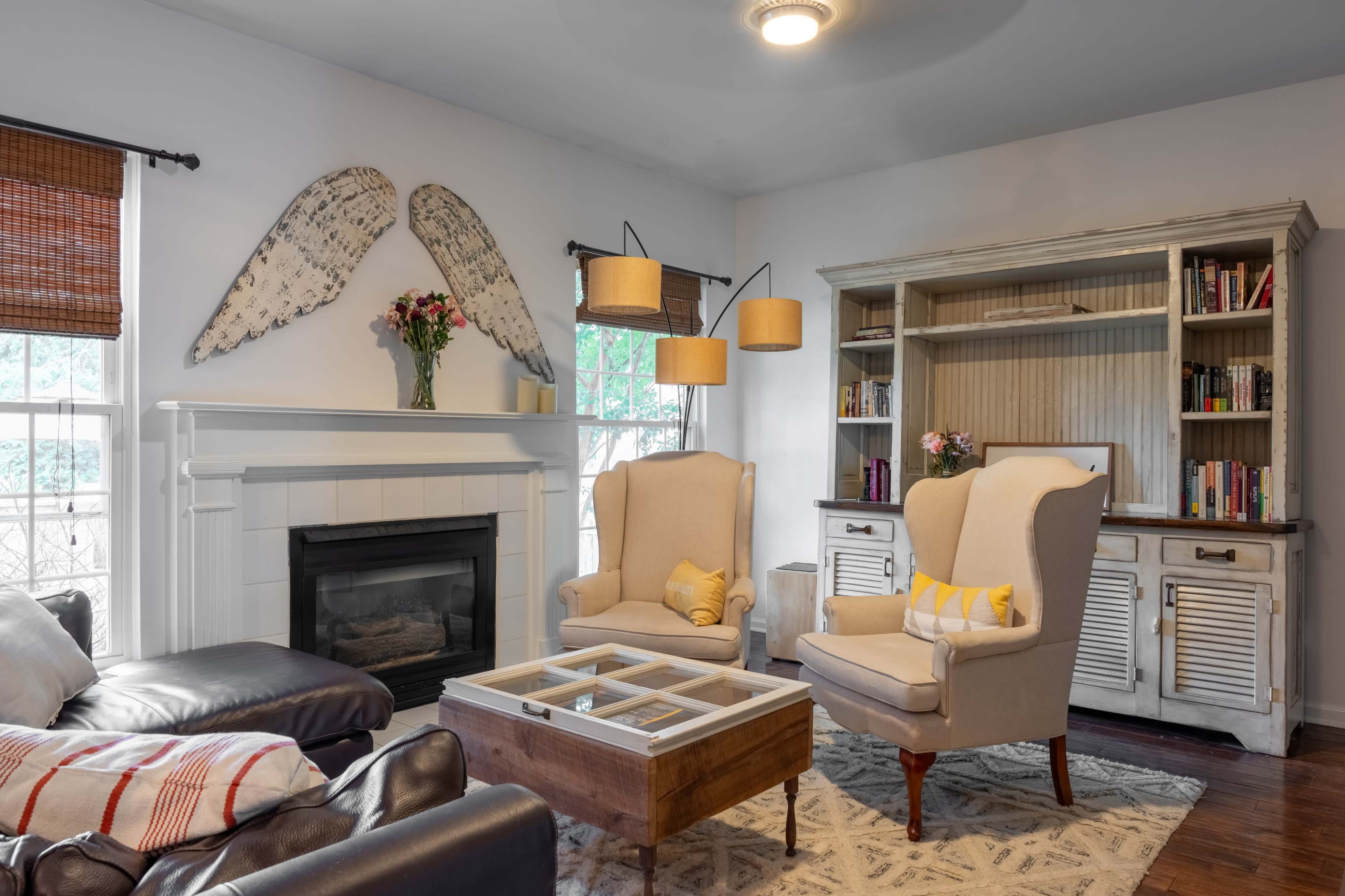 The living room features a fireplace with a white mantel, two armchairs, a coffee table with a glass top, and a bookshelf, all accented by decorative wall pieces and warm lighting.