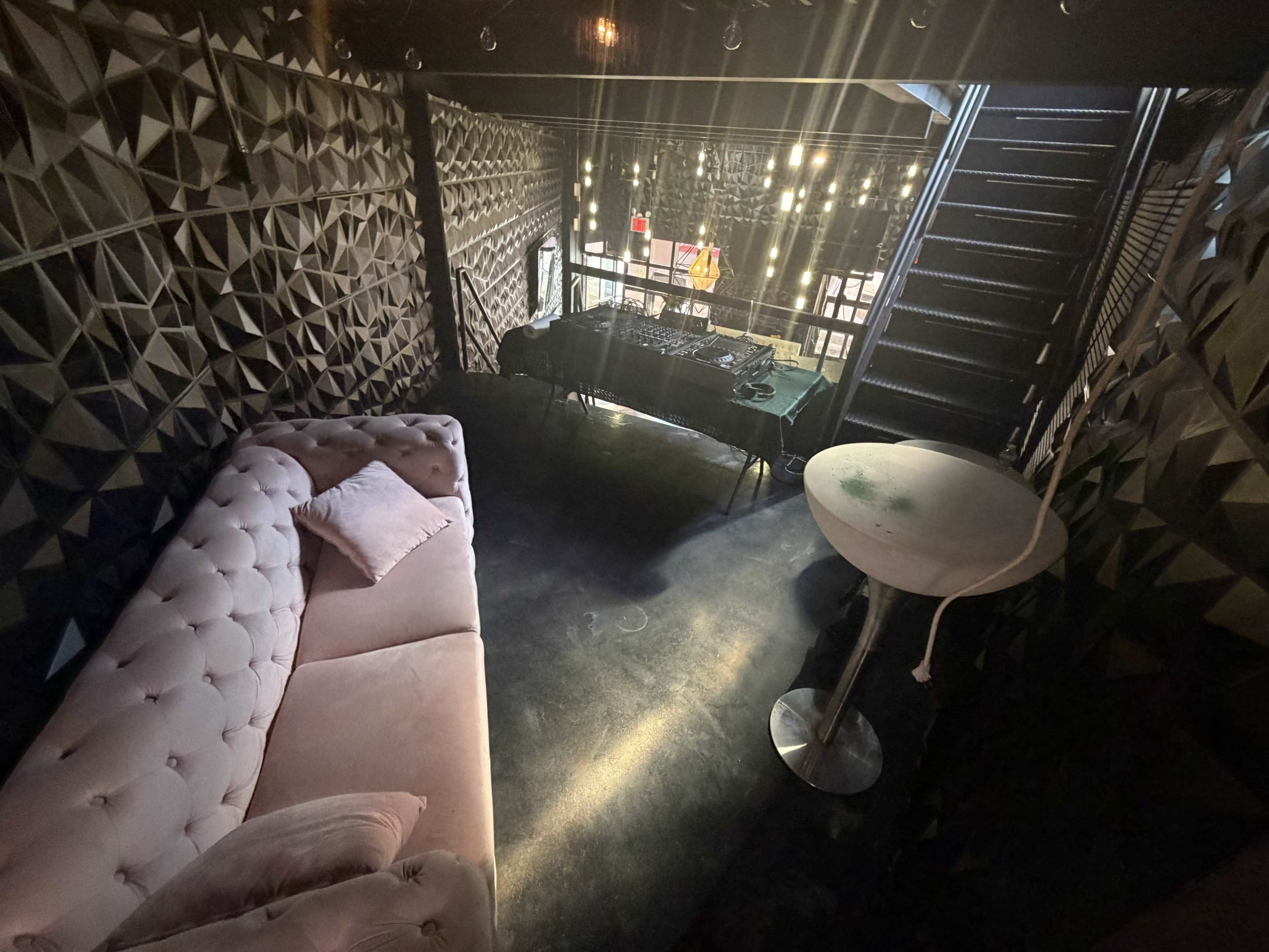 The image shows a dimly lit, modern lounge area with a pink tufted couch, a small white table, and a staircase leading to an upper level.