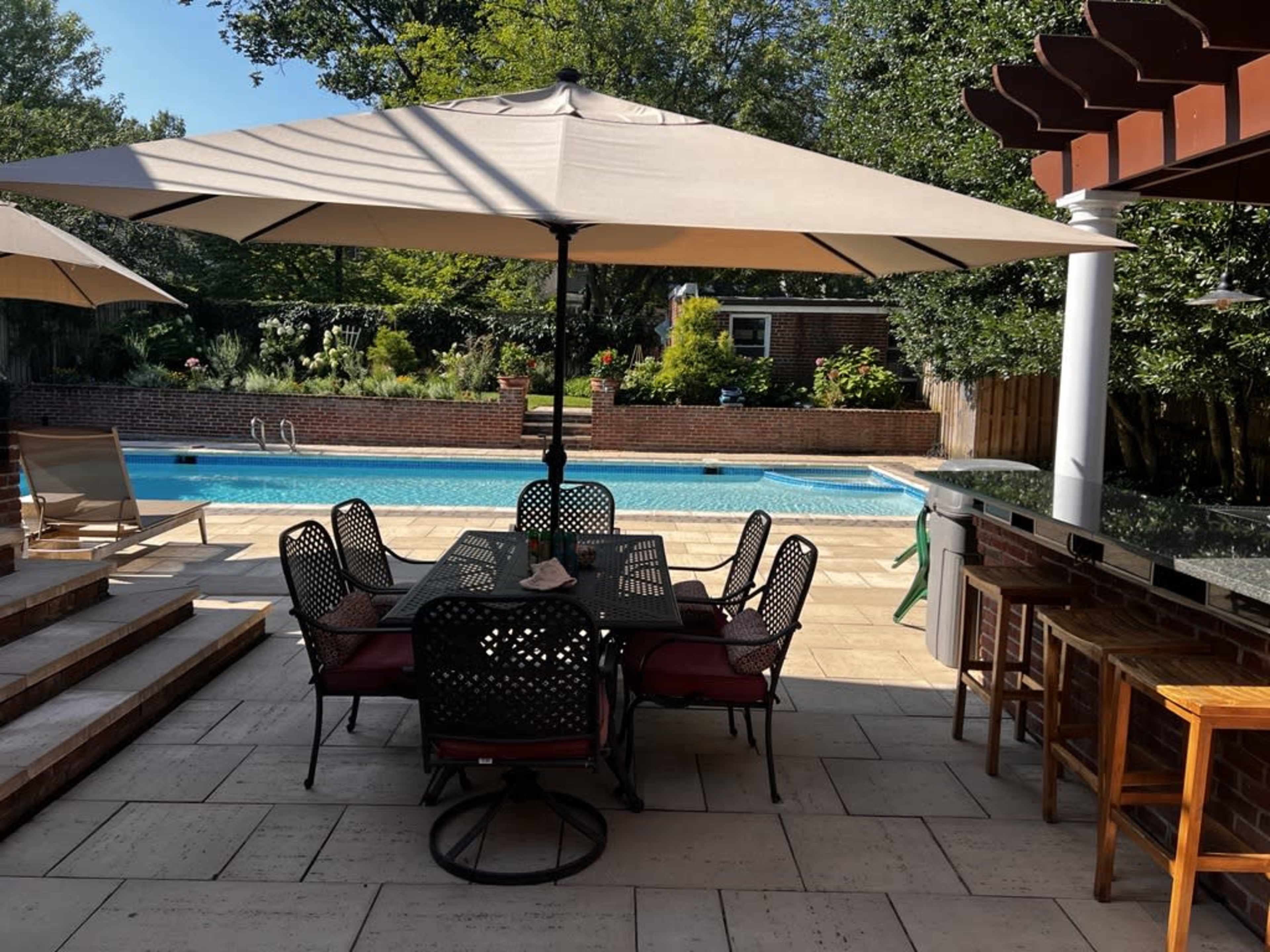 A patio area features a large dining table set with chairs, shaded by an umbrella, alongside a swimming pool surrounded by greenery.