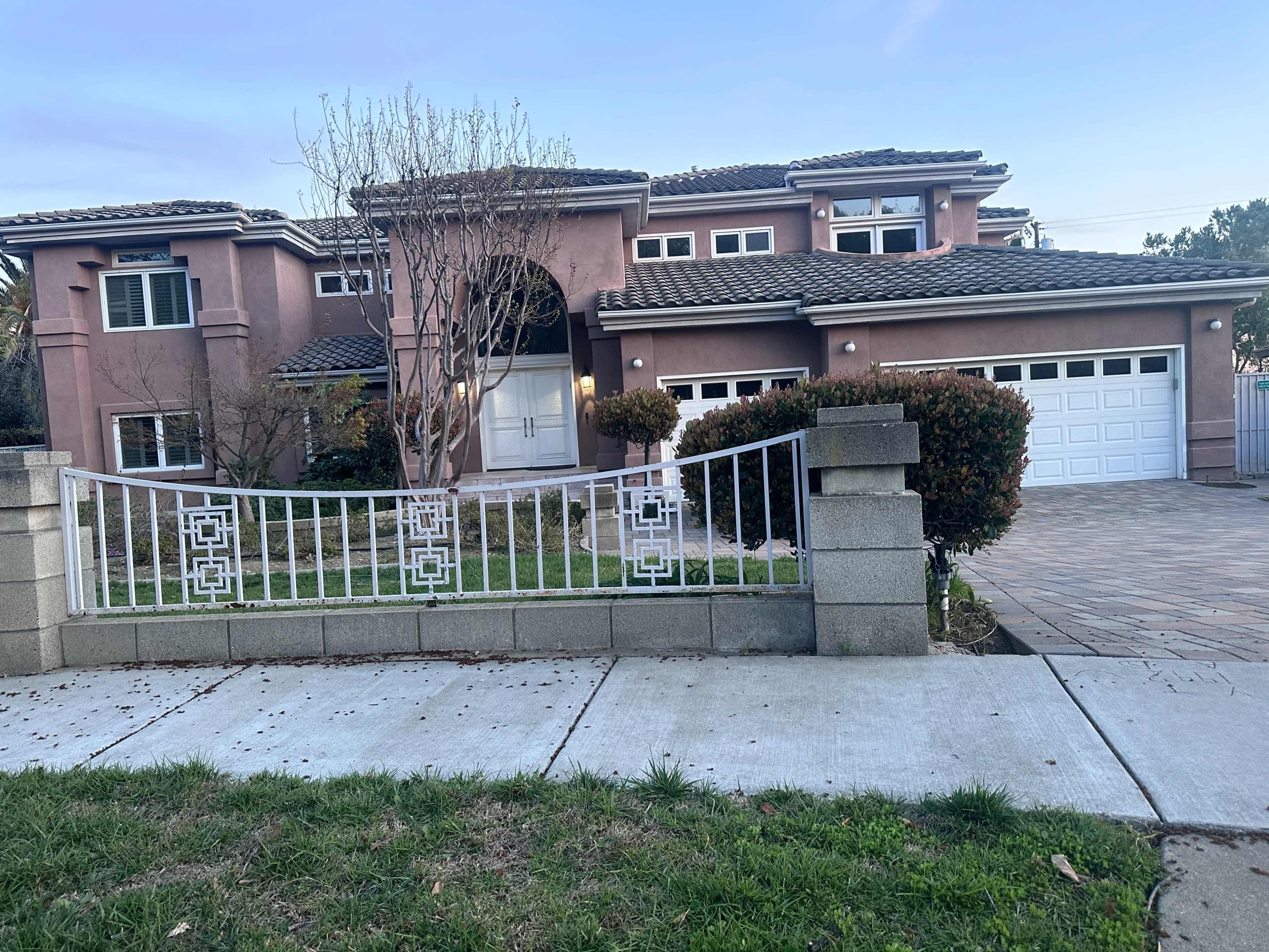 A large two-story house with a pink exterior and a tile roof is set behind a decorative white gate and fence.