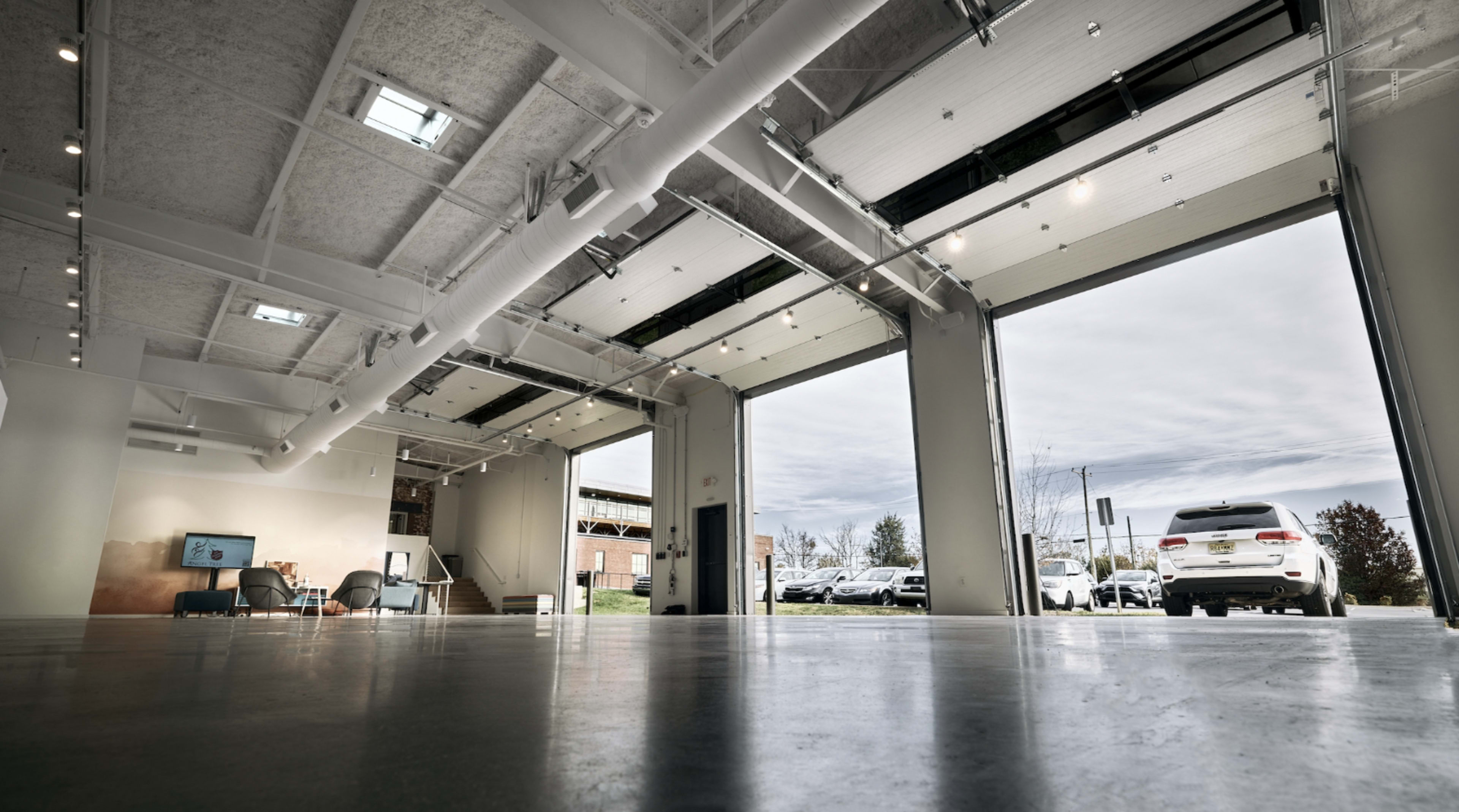 The image shows a spacious, modern garage with large open doors allowing a view of parked cars outside.