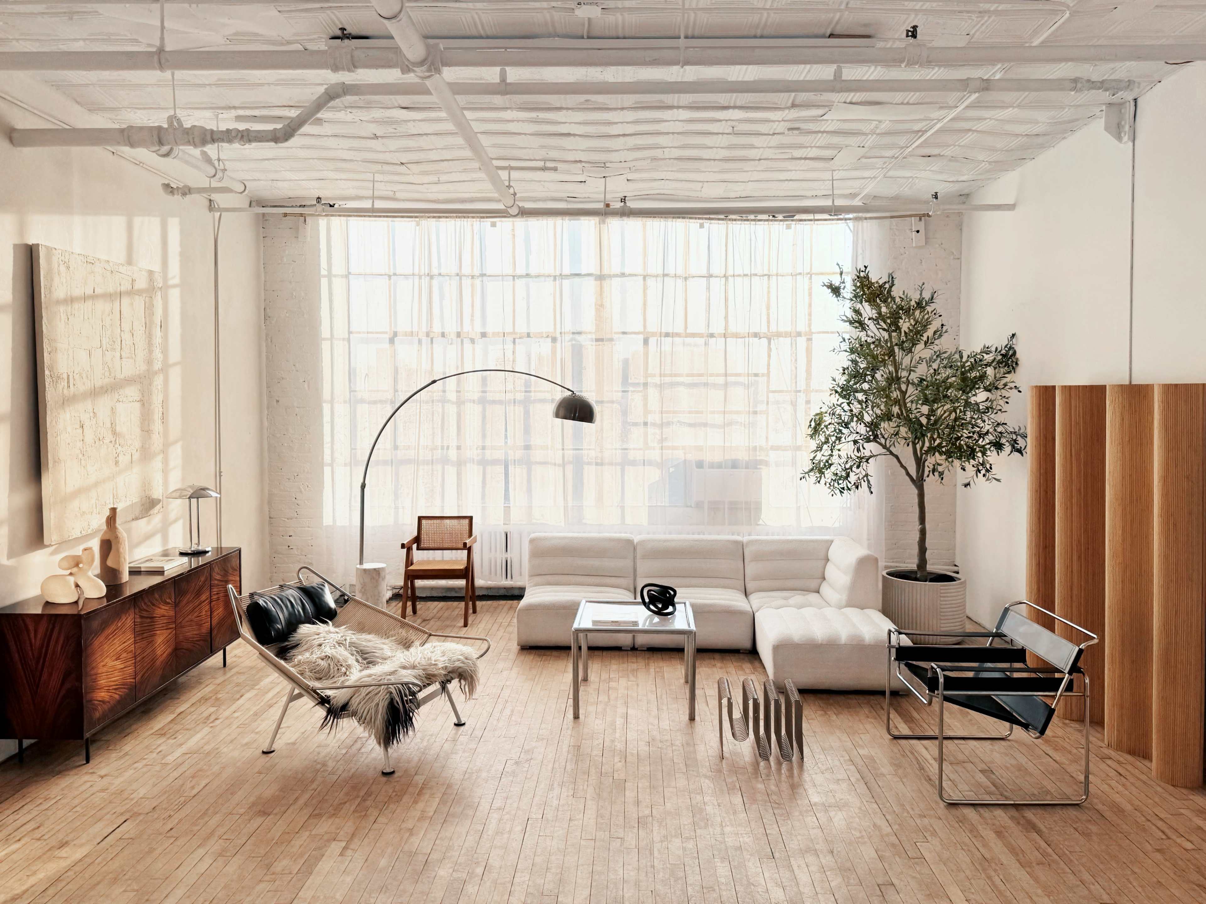 The image shows a modern living room with a white sofa, a black lounge chair, a wooden sideboard, and large windows allowing natural light to fill the space.