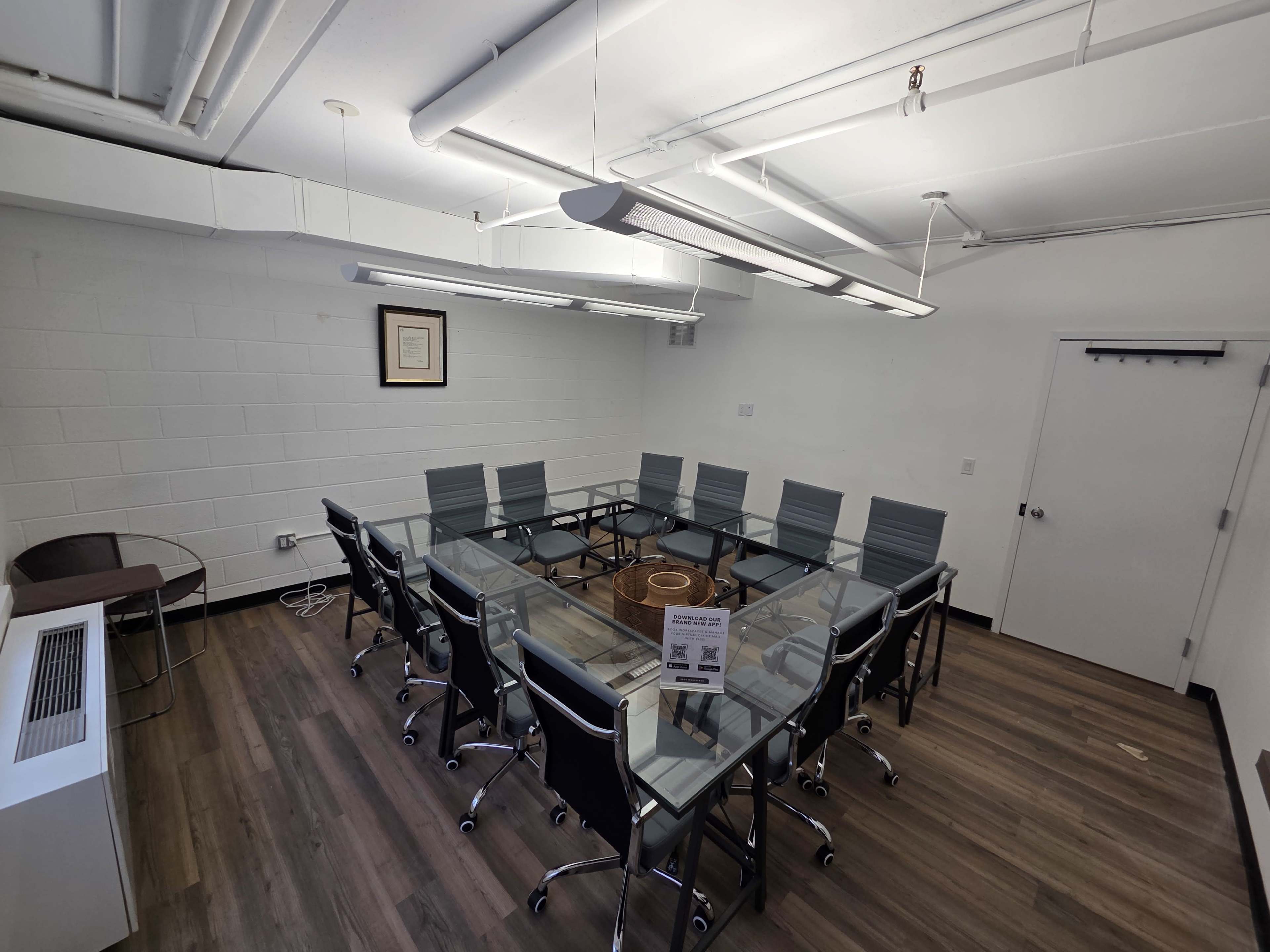 The image shows a modern conference room with a large glass table surrounded by gray office chairs, illuminated by overhead lights.