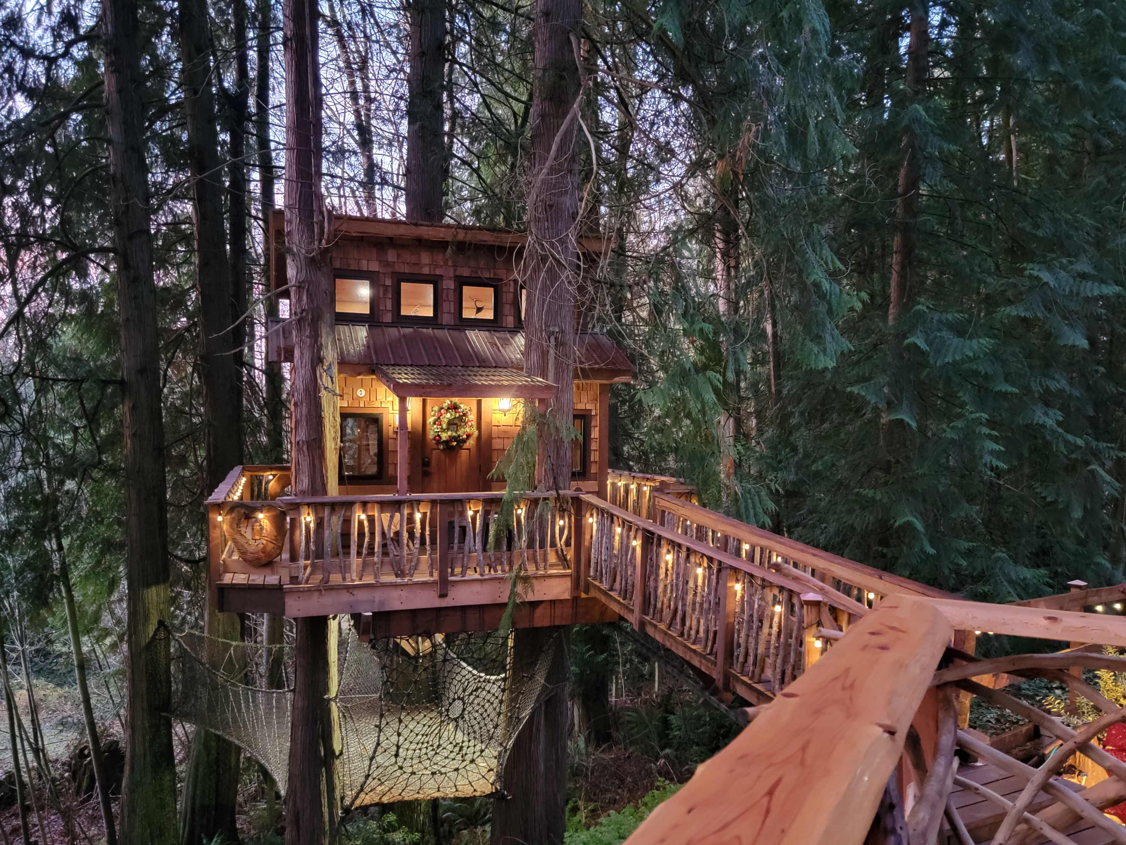 A wooden treehouse with a balcony and lights is elevated among tall trees in a forested area.
