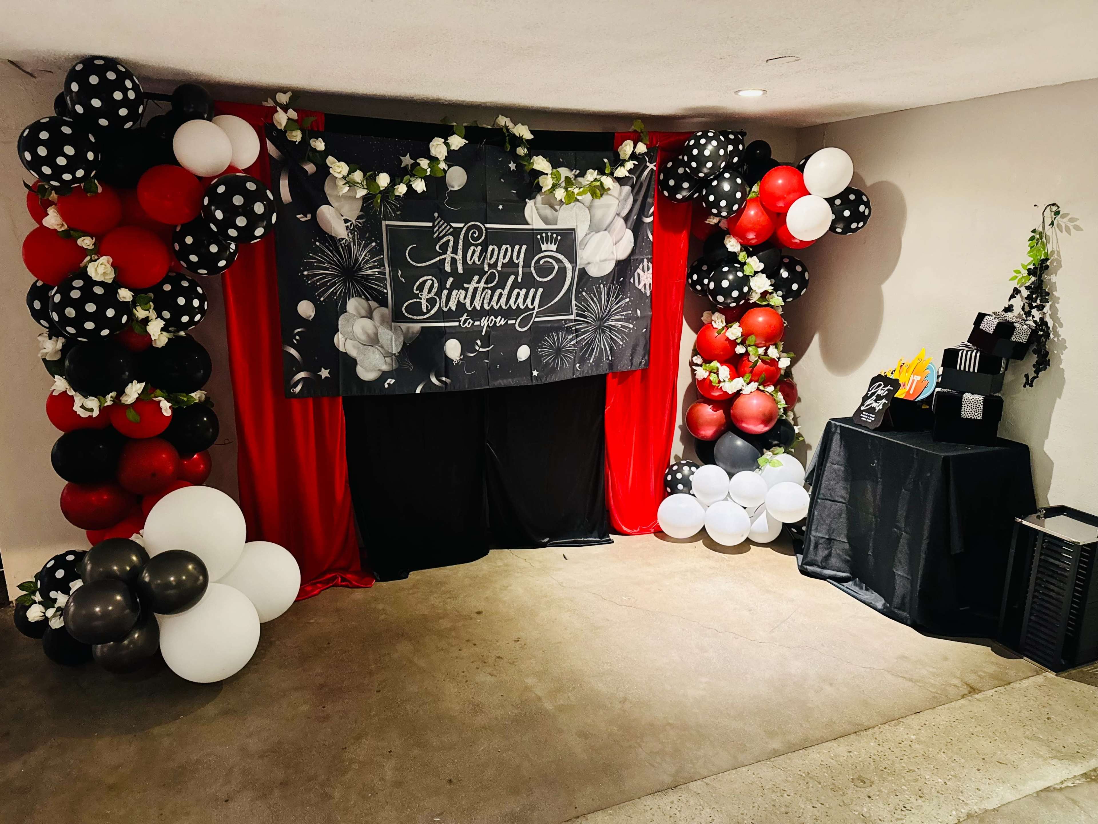 The image shows a birthday celebration setup featuring a decorative backdrop with the text "Happy Birthday," surrounded by colorful balloons in red, black, white, and silver, along with a table displaying party supplies.
