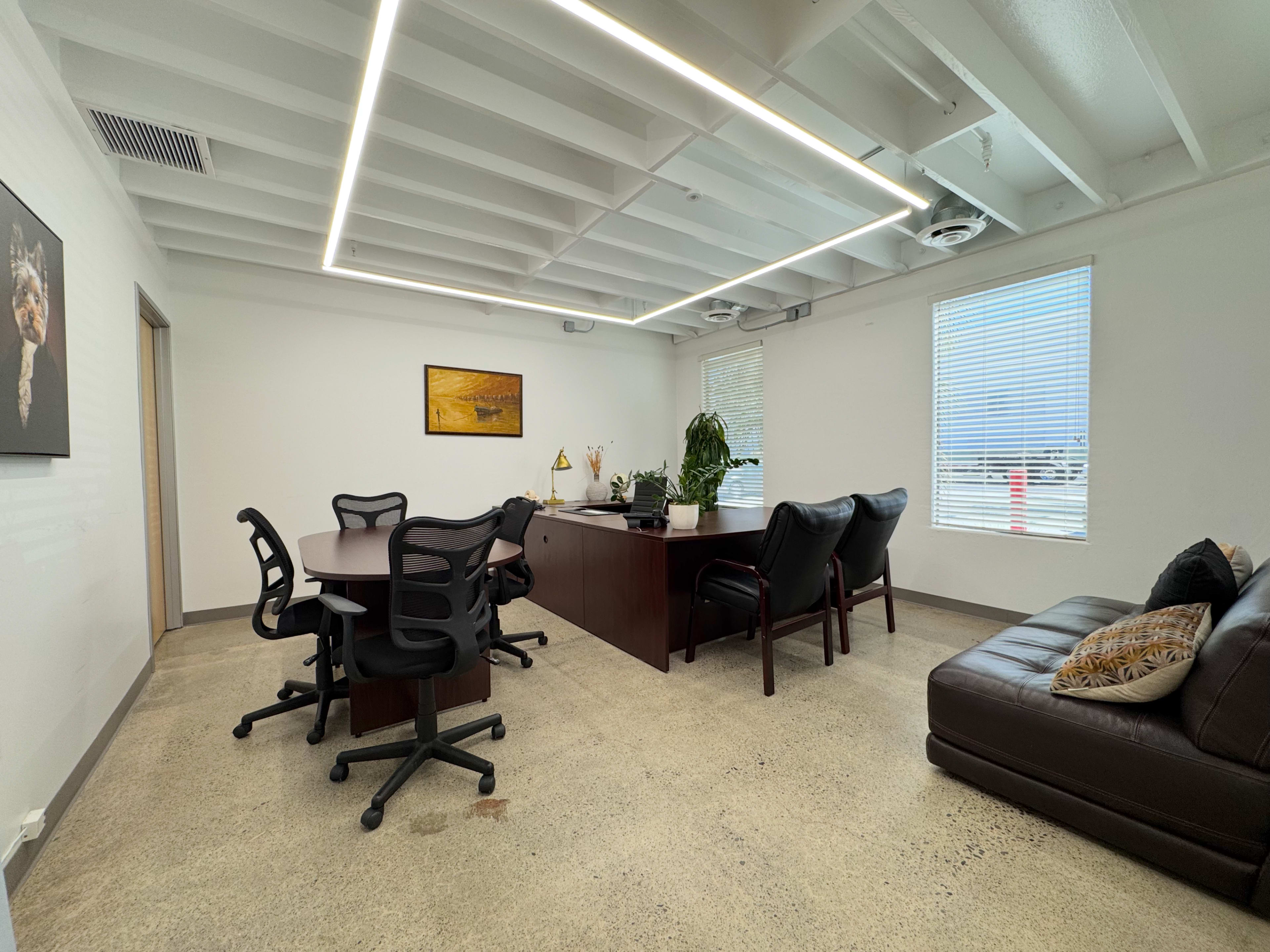 The image shows a modern office space with a conference table, several chairs, an executive desk, and large windows allowing natural light to enter.
