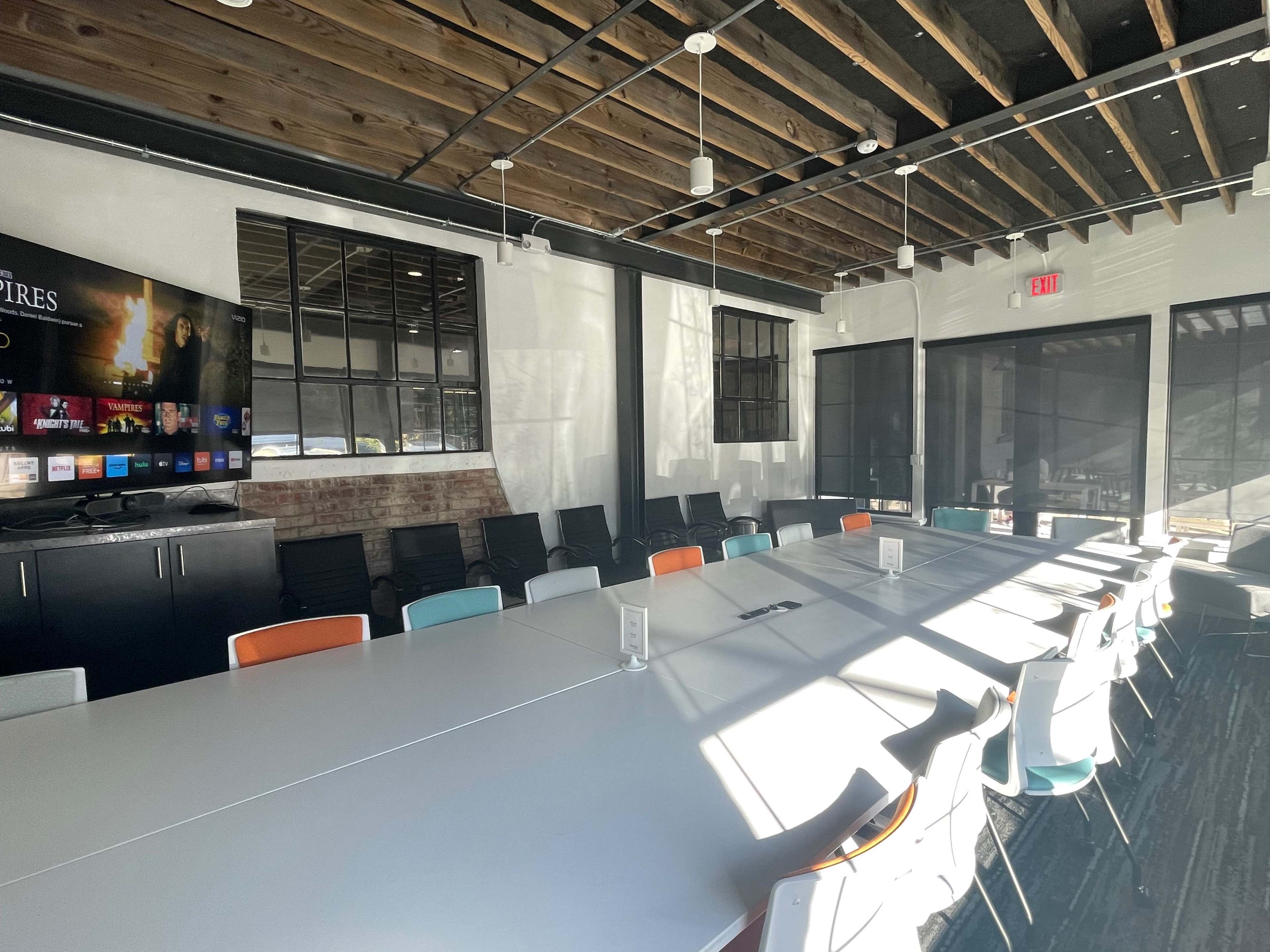 A modern conference room features a long rectangular table surrounded by chairs, with large windows and a wall-mounted screen displaying a menu.