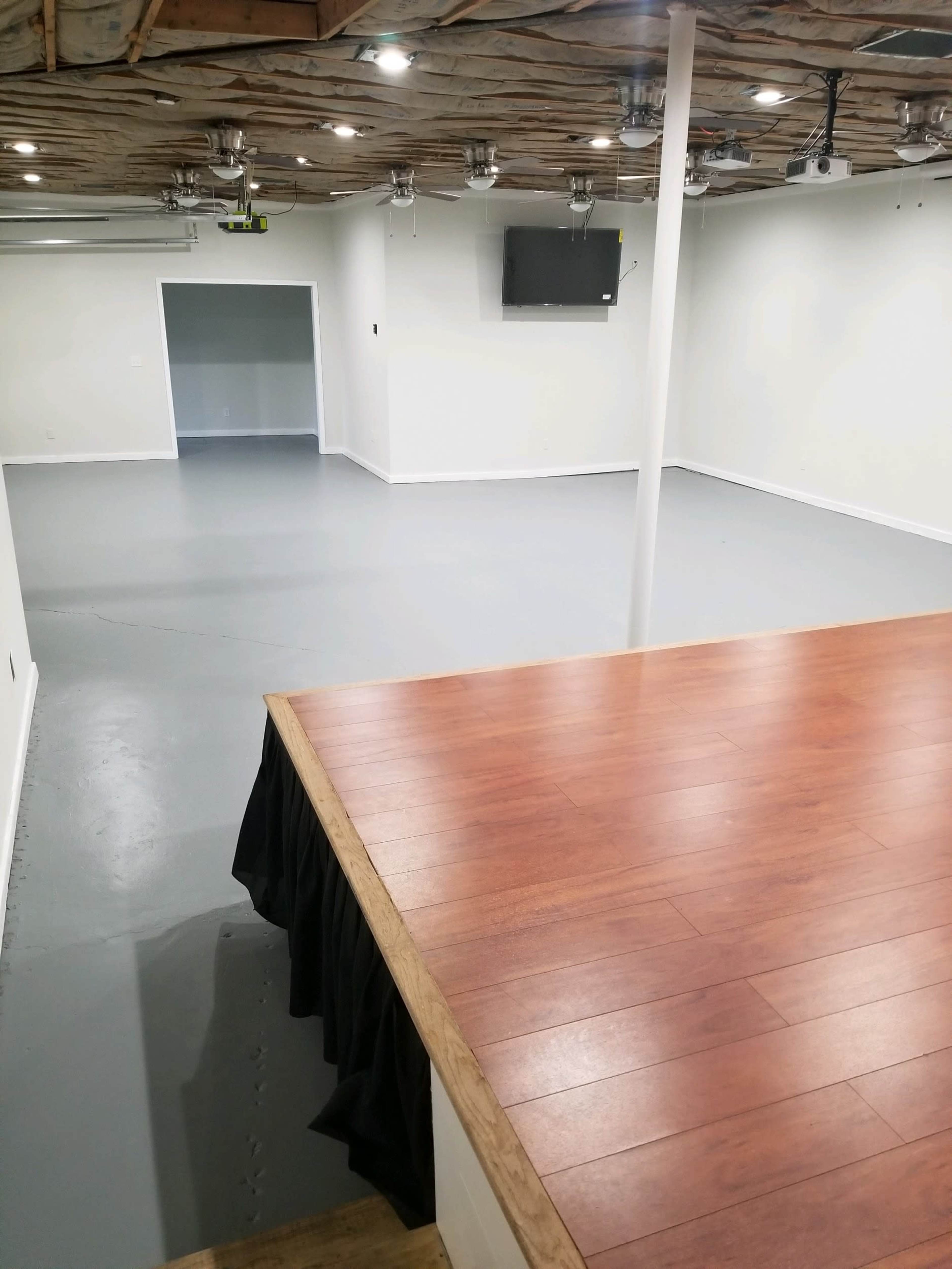An empty room with a wooden stage in the foreground and light gray flooring, featuring a television mounted on the wall.
