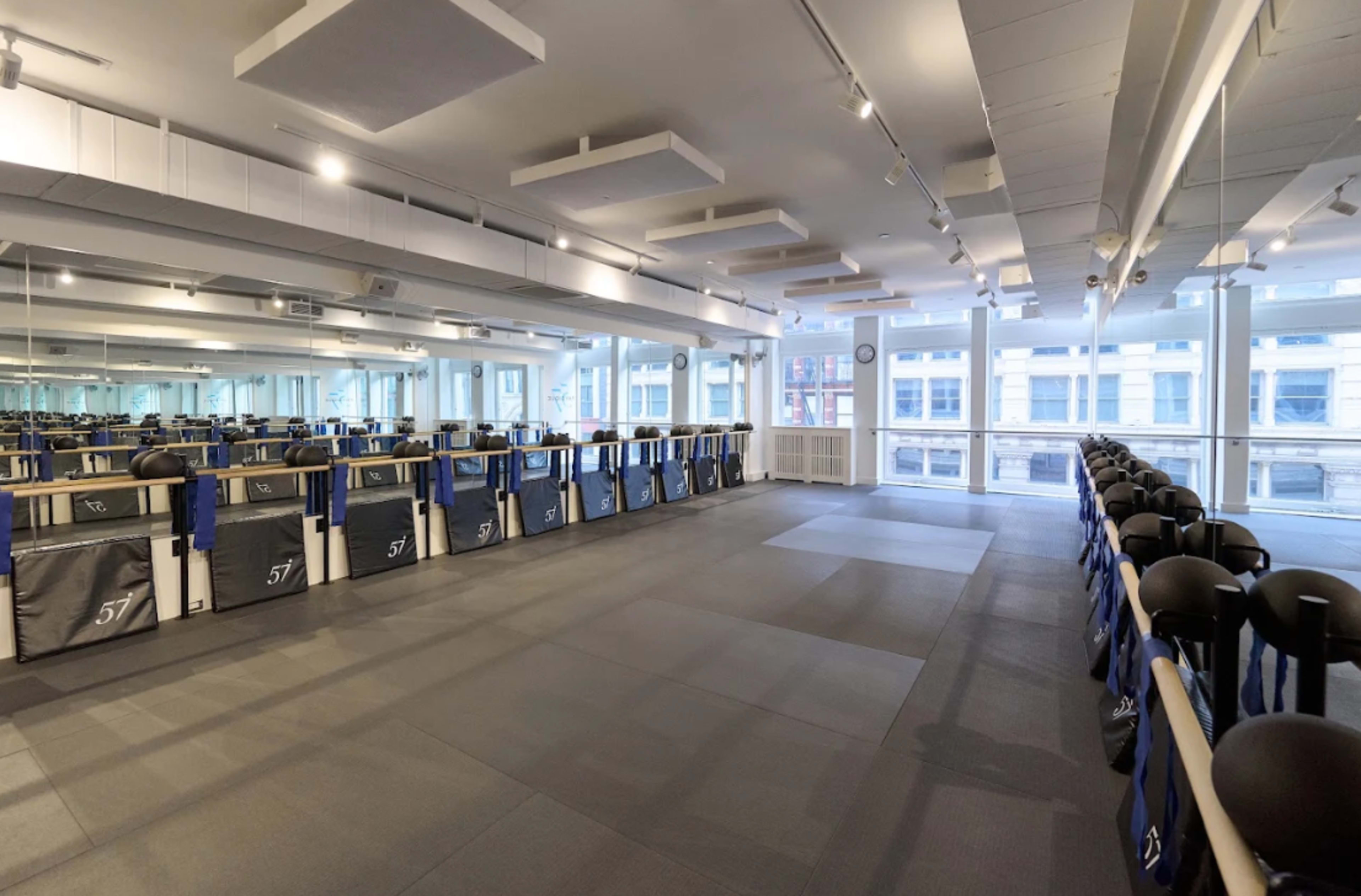 The image shows an empty gym studio with mirrored walls, a wooden barre, and gray flooring, illuminated by natural light from large windows.