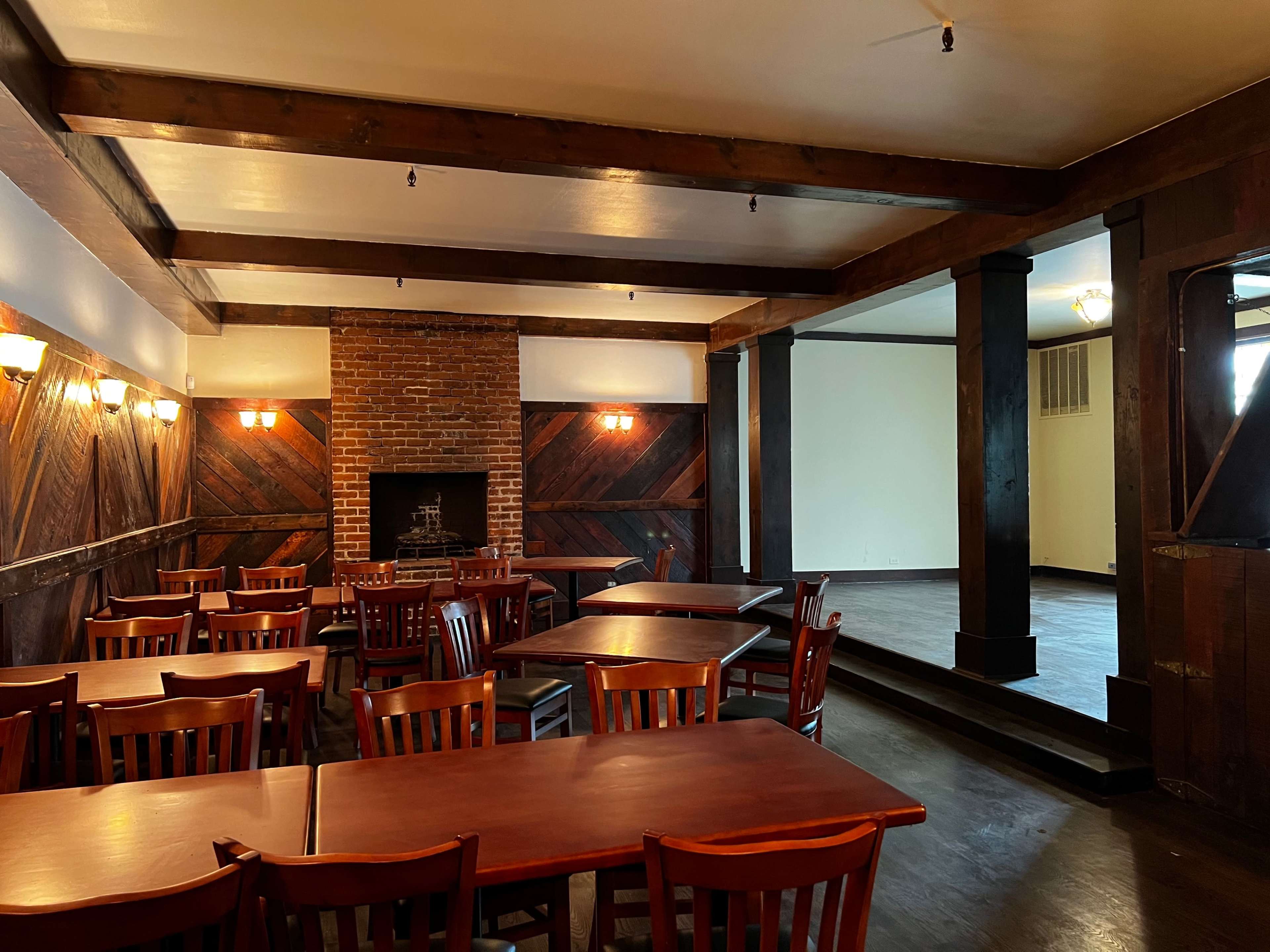 A restaurant interior with wooden tables and chairs arranged around a brick fireplace, featuring exposed beams and a separate empty stage area.