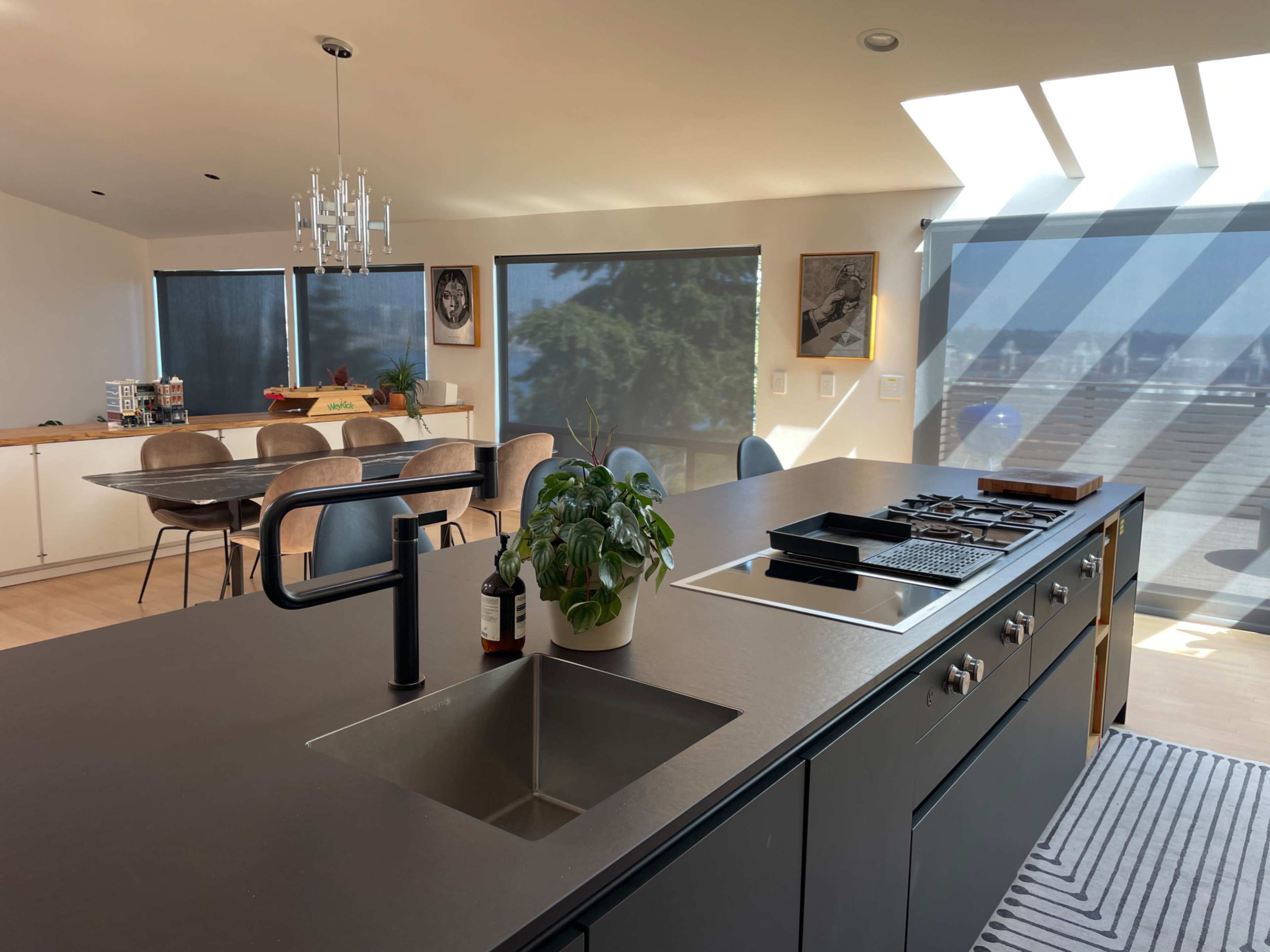 A modern kitchen with a central island featuring a sink and a cooktop, overlooking a bright dining area through large windows.