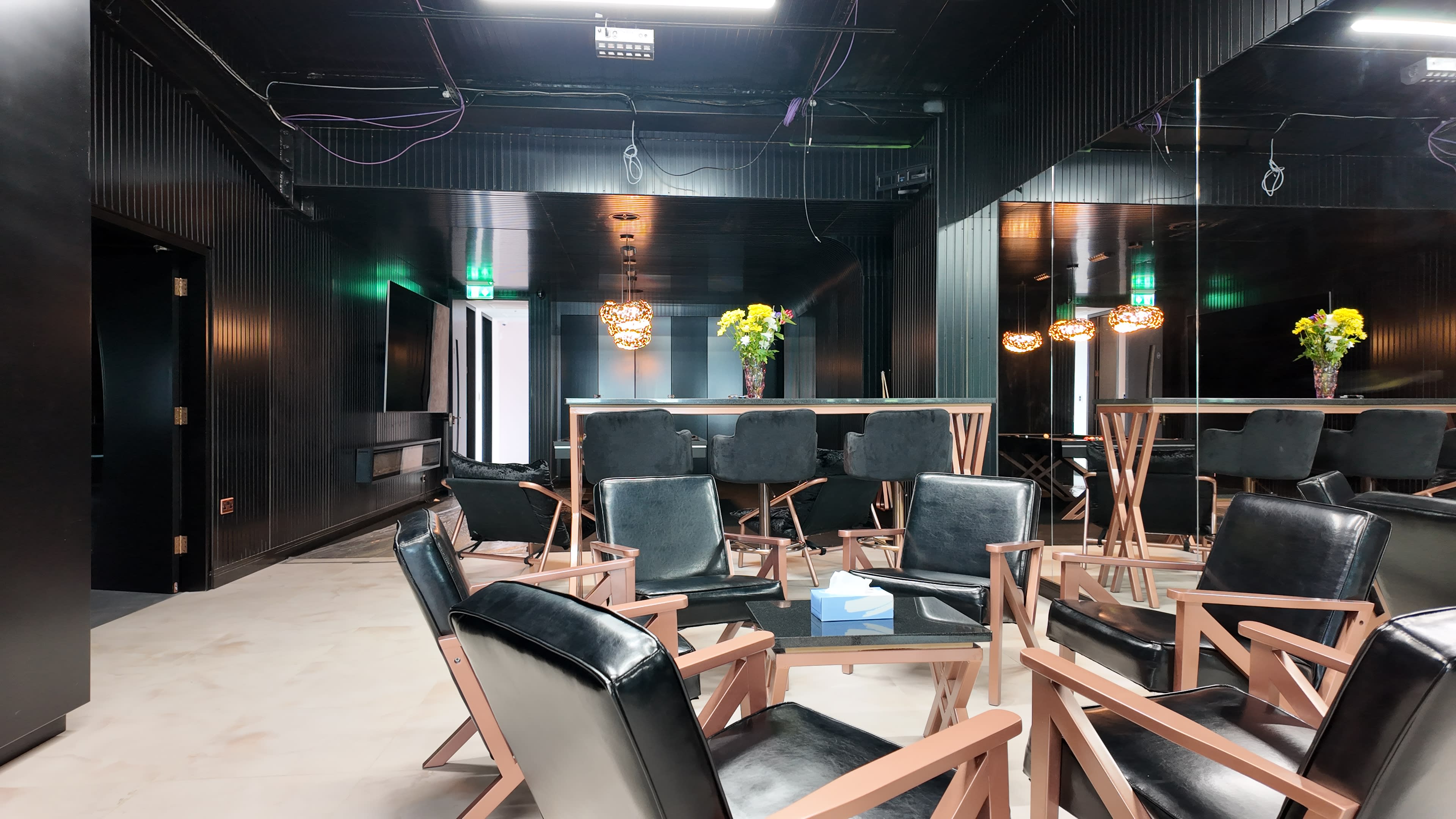 The image shows a modern, dimly lit lounge area with black walls, wooden chairs, and a mirrored backdrop, featuring a table with a small stack of tissues and a vase of flowers.