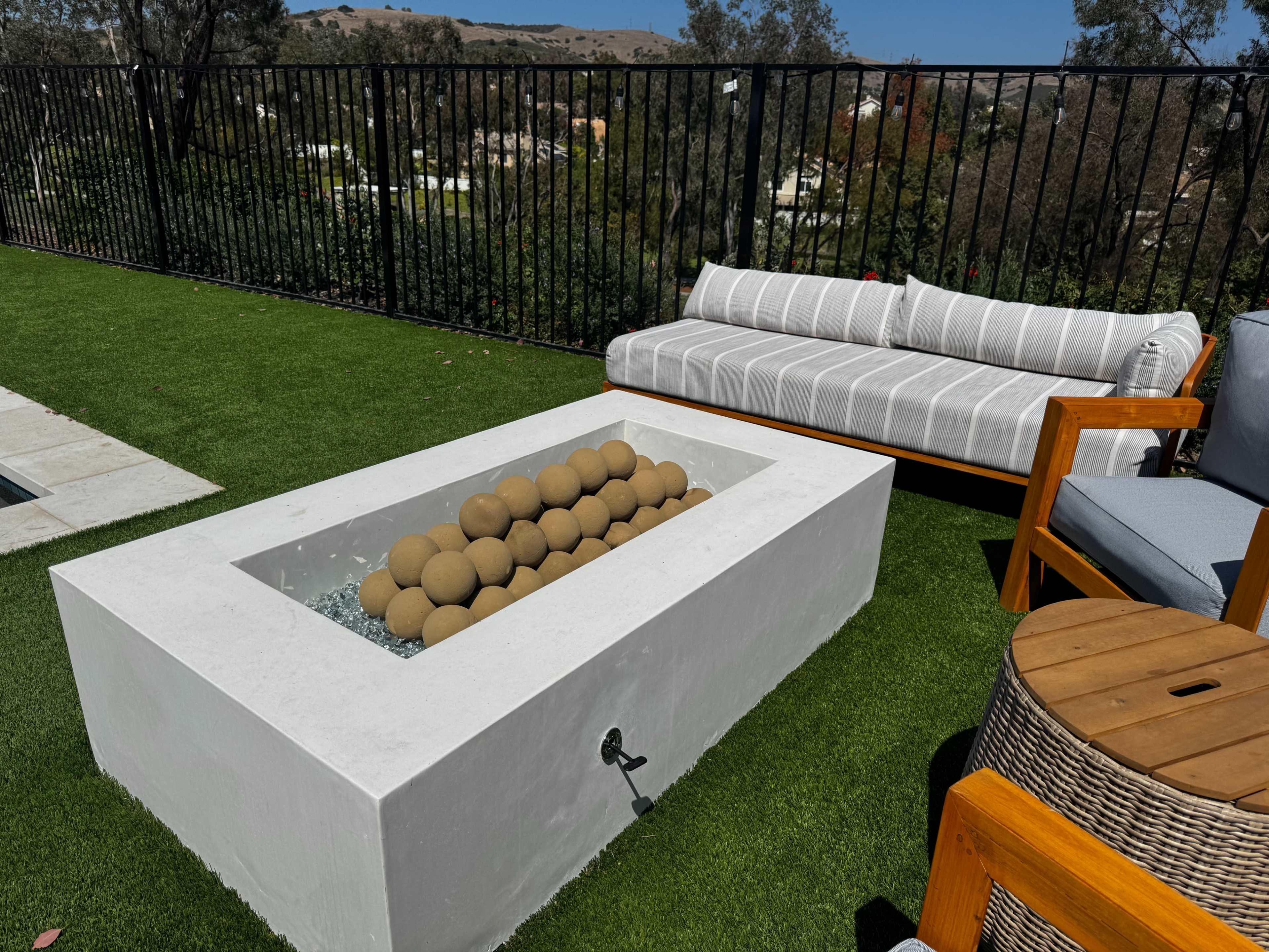 The image shows a modern outdoor space featuring a white fire pit with decorative stones, a gray and white striped sofa, and a wooden chair, all set on synthetic grass.