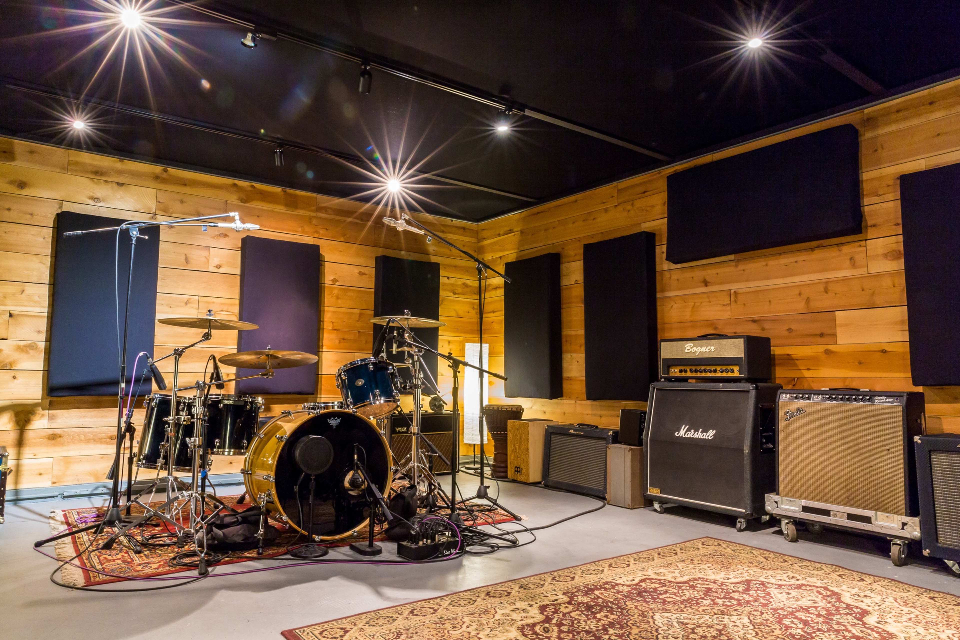 A recording studio features a drum set, microphones, and amplifiers against wooden paneling and soundproofing.
