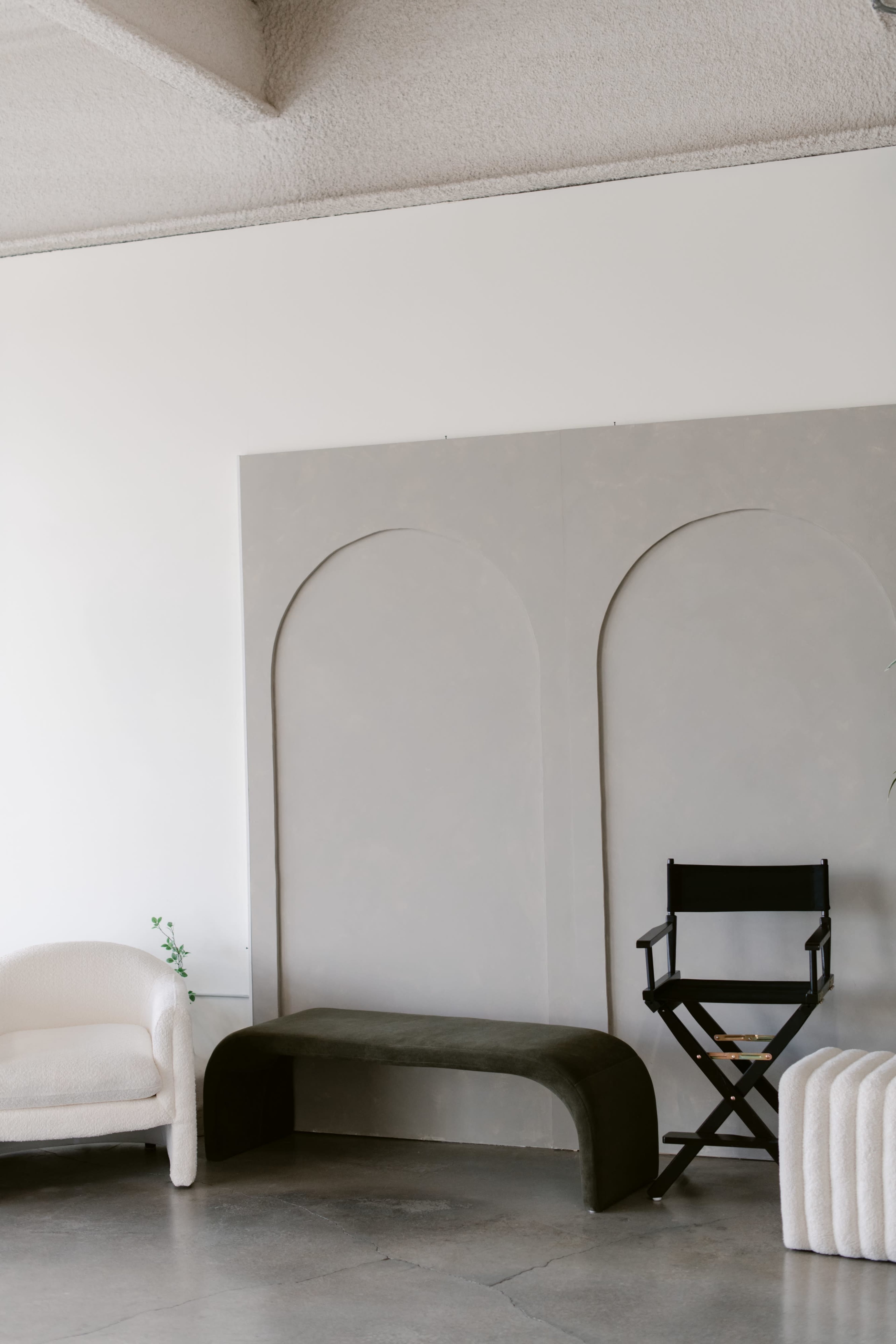 The scene features a minimalistic interior with a textured gray wall, a curved green bench, a black director's chair, and a white upholstered sofa.