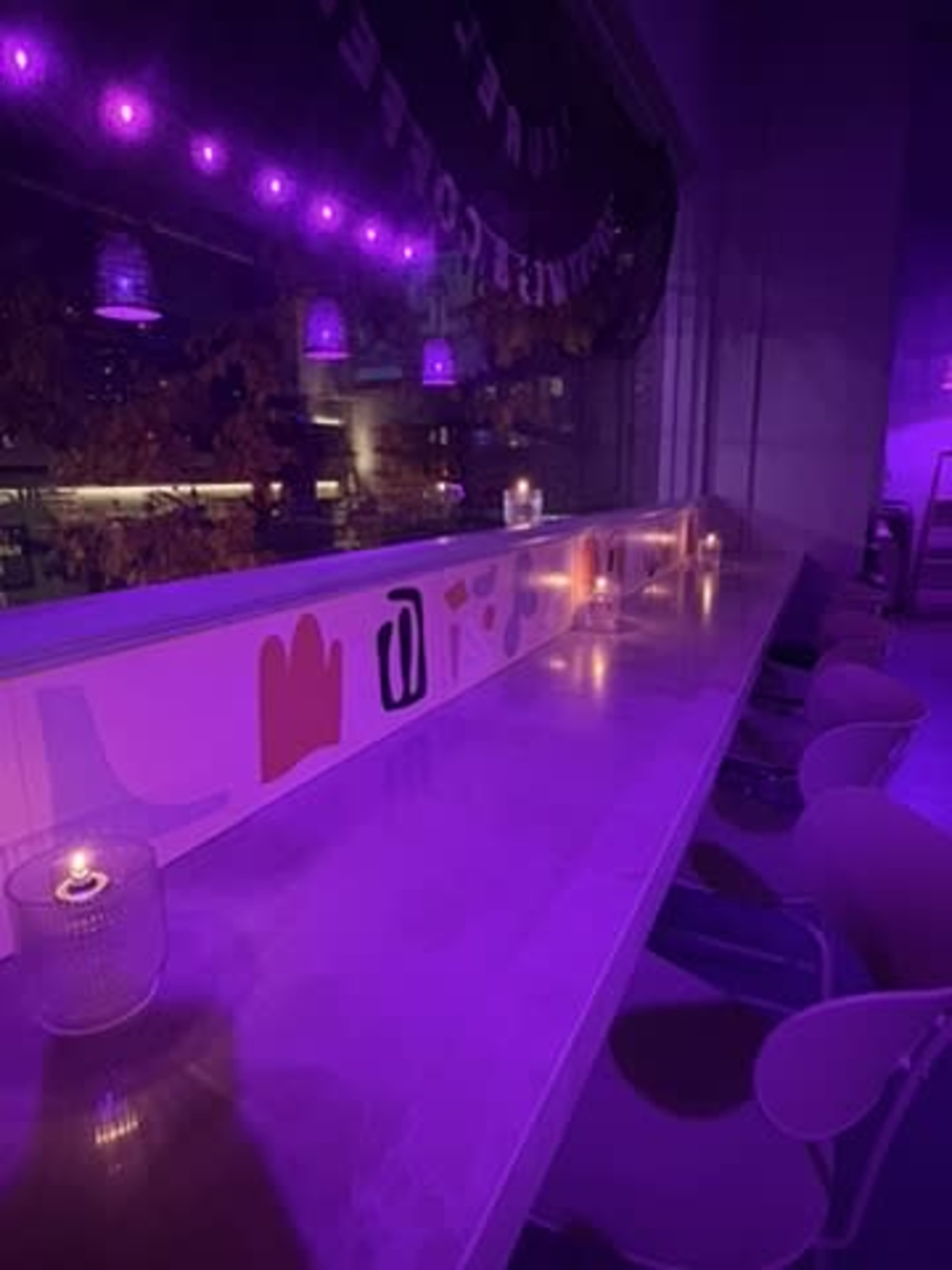 A dimly lit café features a series of clear candles on a long, glossy table surrounded by modern chairs, with colorful artwork displayed on the wall near large windows.