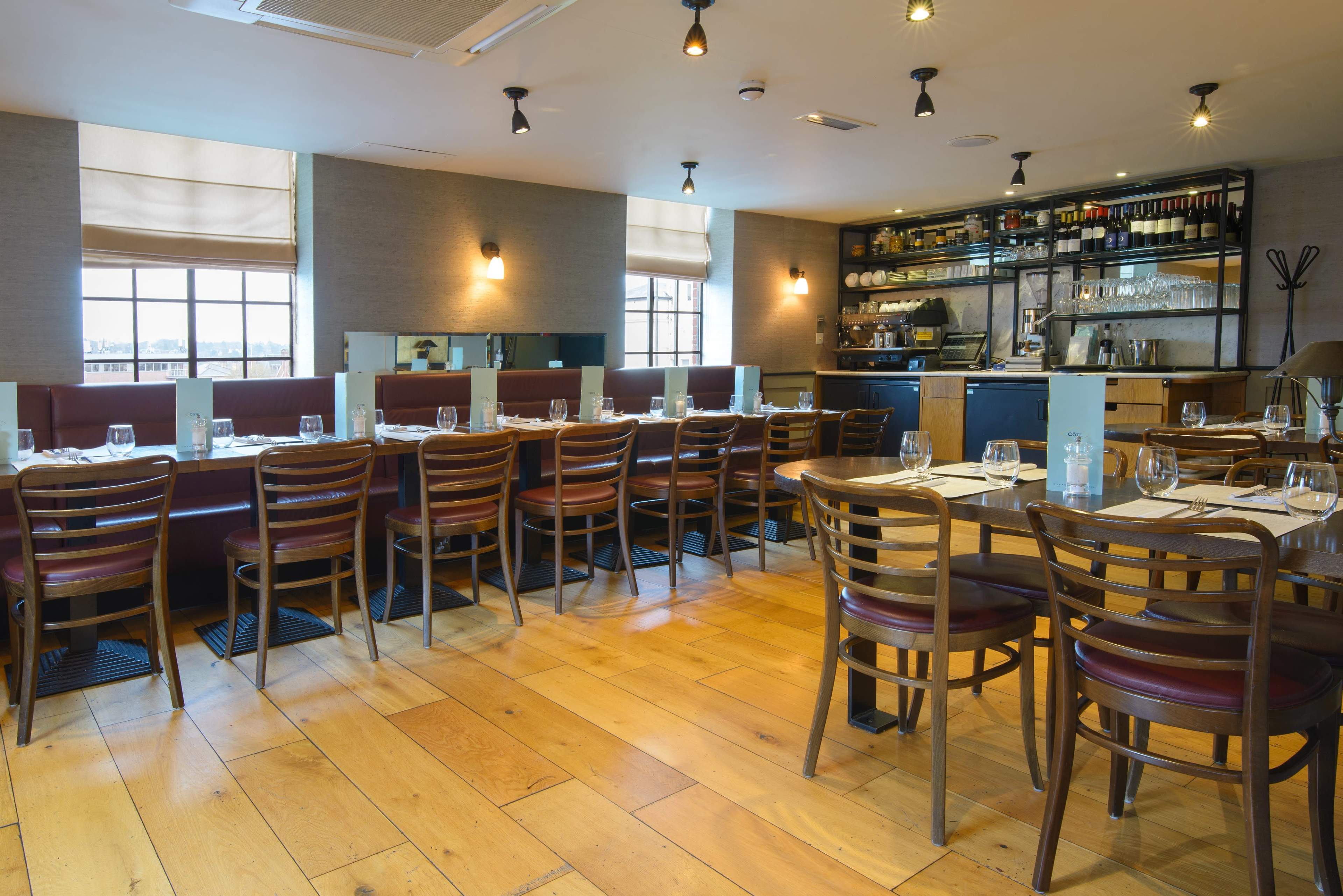 The image shows a restaurant interior with wooden floors, rows of tables, and a bar area in the background.
