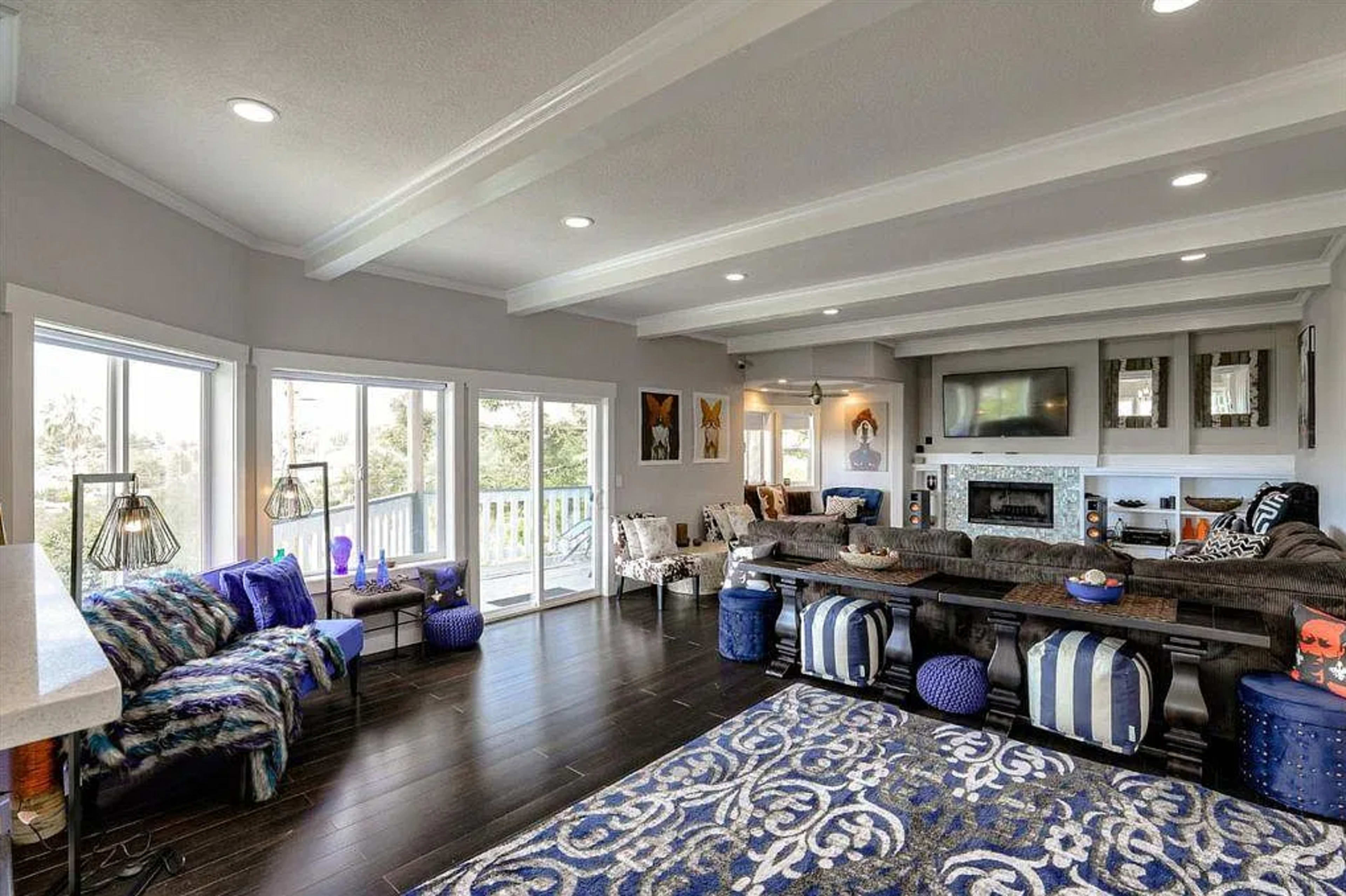 The image shows a spacious living room with large windows, a gray couch, decorative blue poufs, and a patterned area rug.