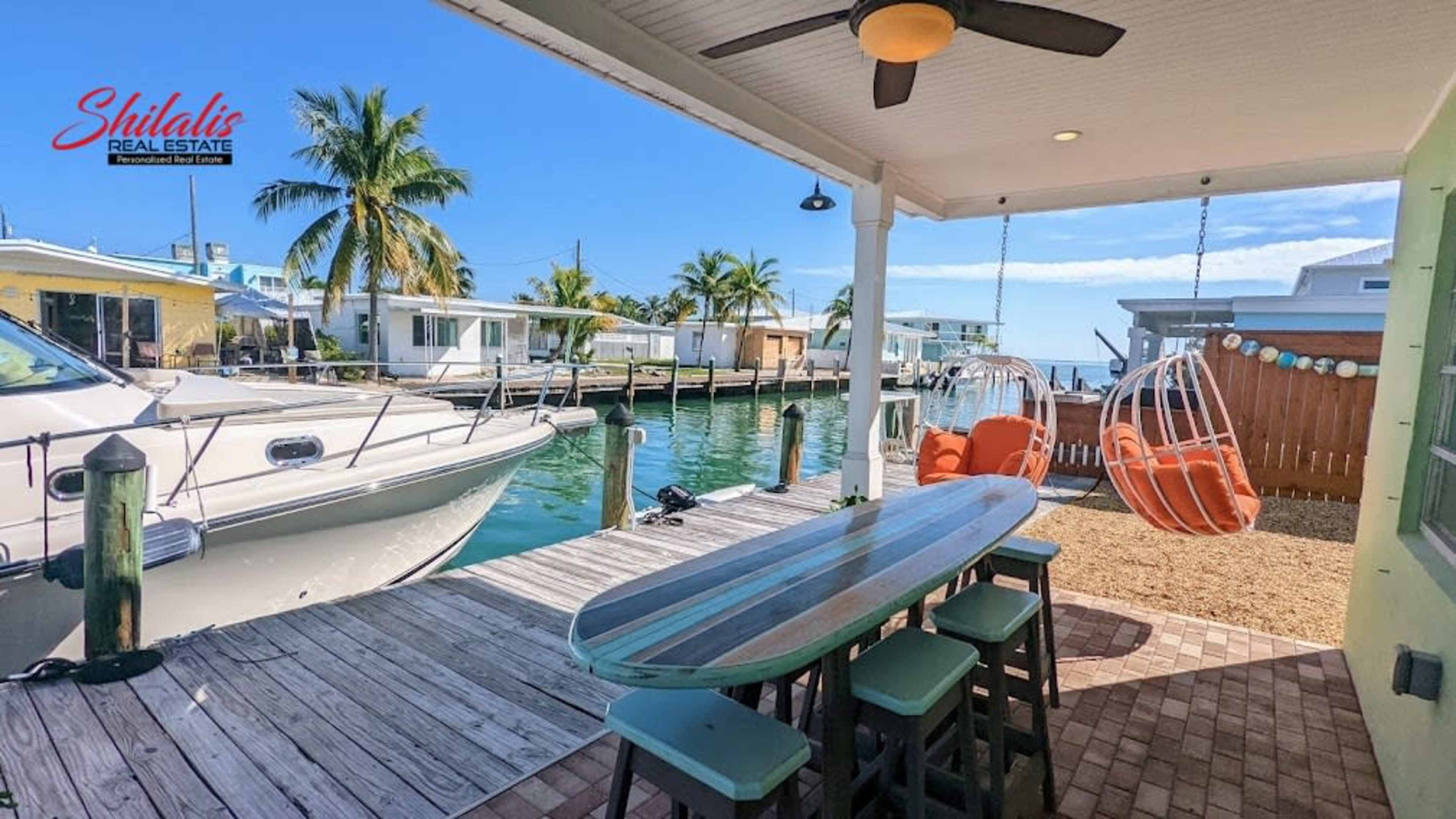 A waterfront patio features a long table and two hanging chairs beside a boat docked in a canal lined with palm trees and houses.
