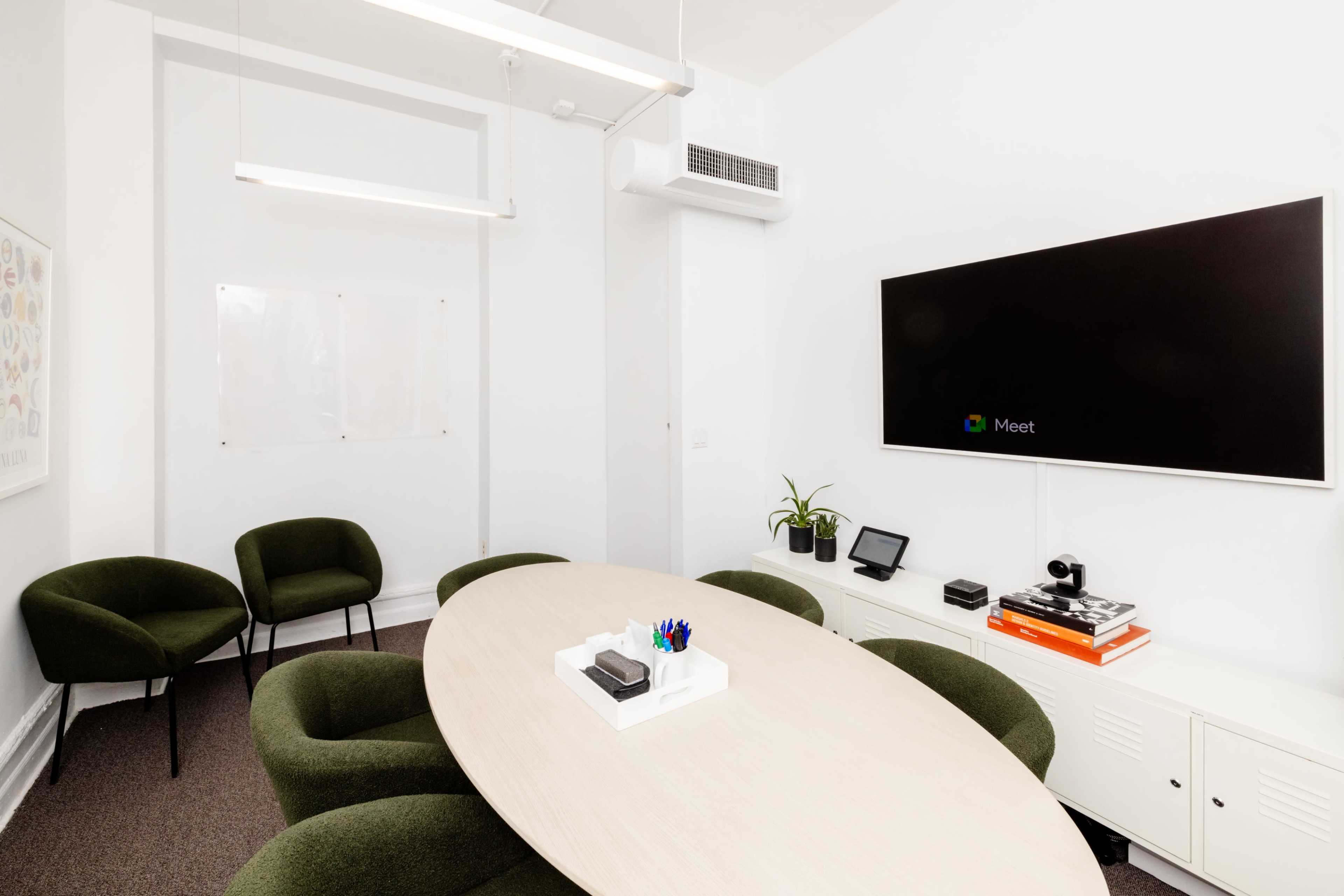 A modern meeting room features a large oval table surrounded by green chairs, with a mounted screen displaying a video conferencing interface.