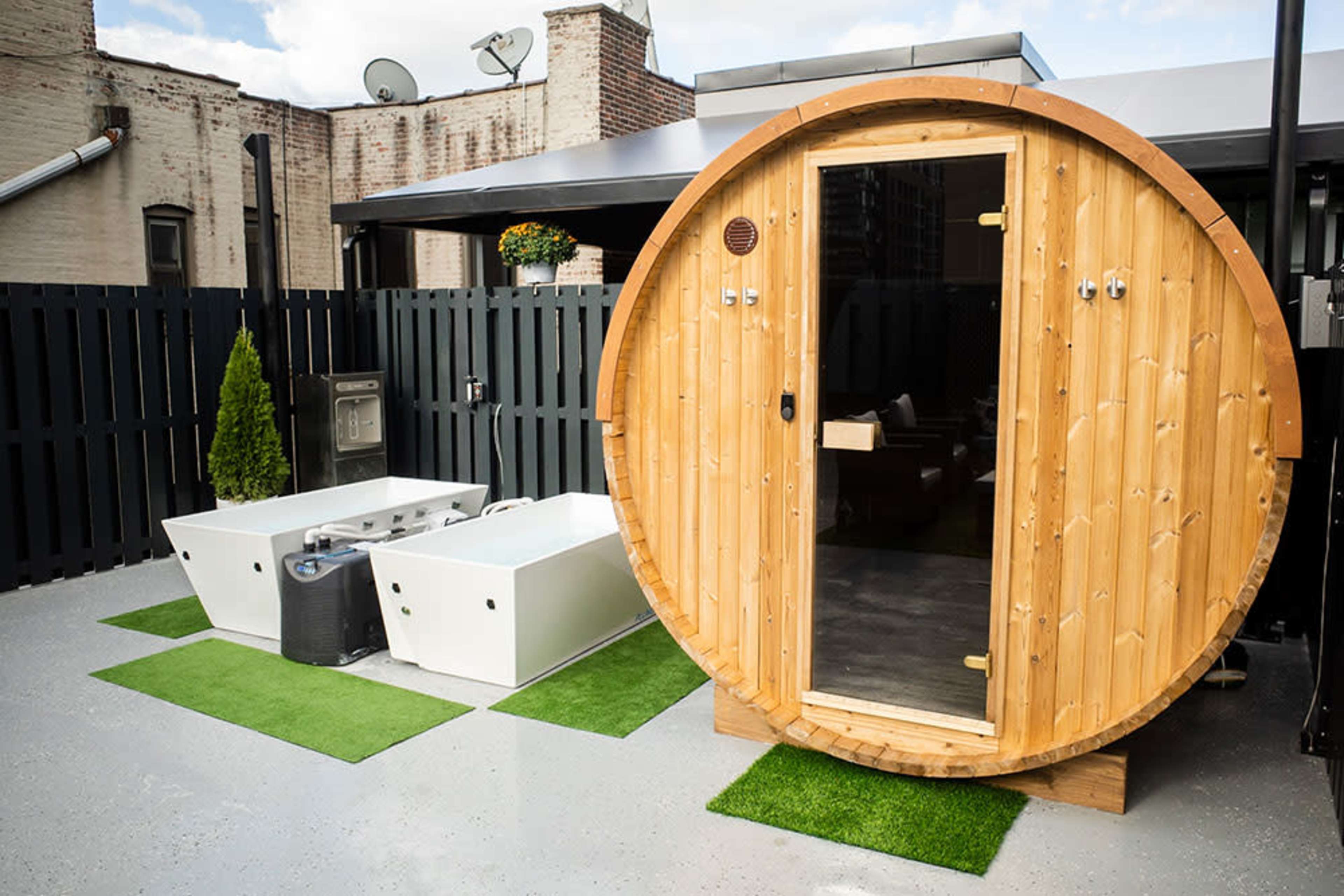 A wooden barrel sauna next to two white hot tubs, set on a grassy area with a dark fence in the background.