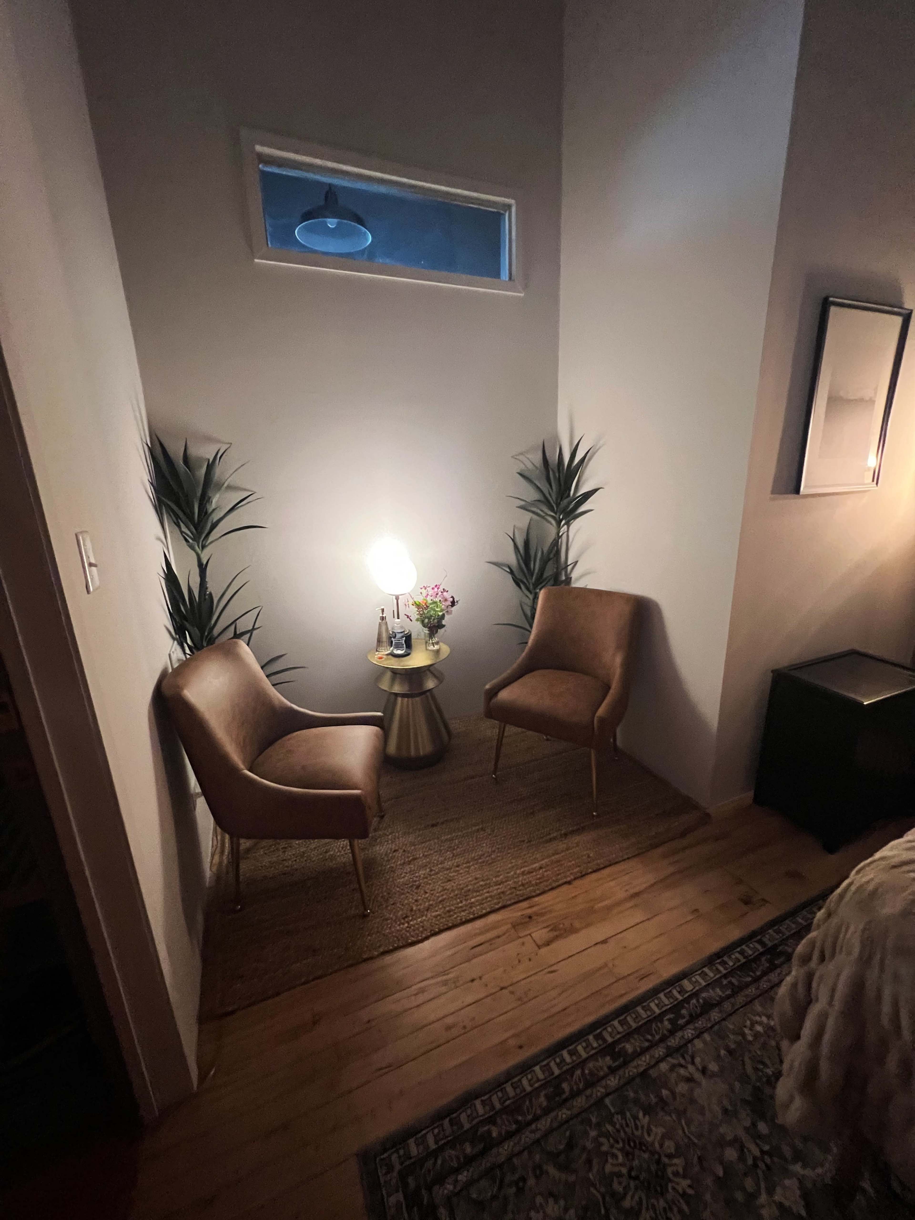 The image shows a small corner of a room featuring two brown chairs, a side table with a lamp and flowers, and two green plants against a light wall.