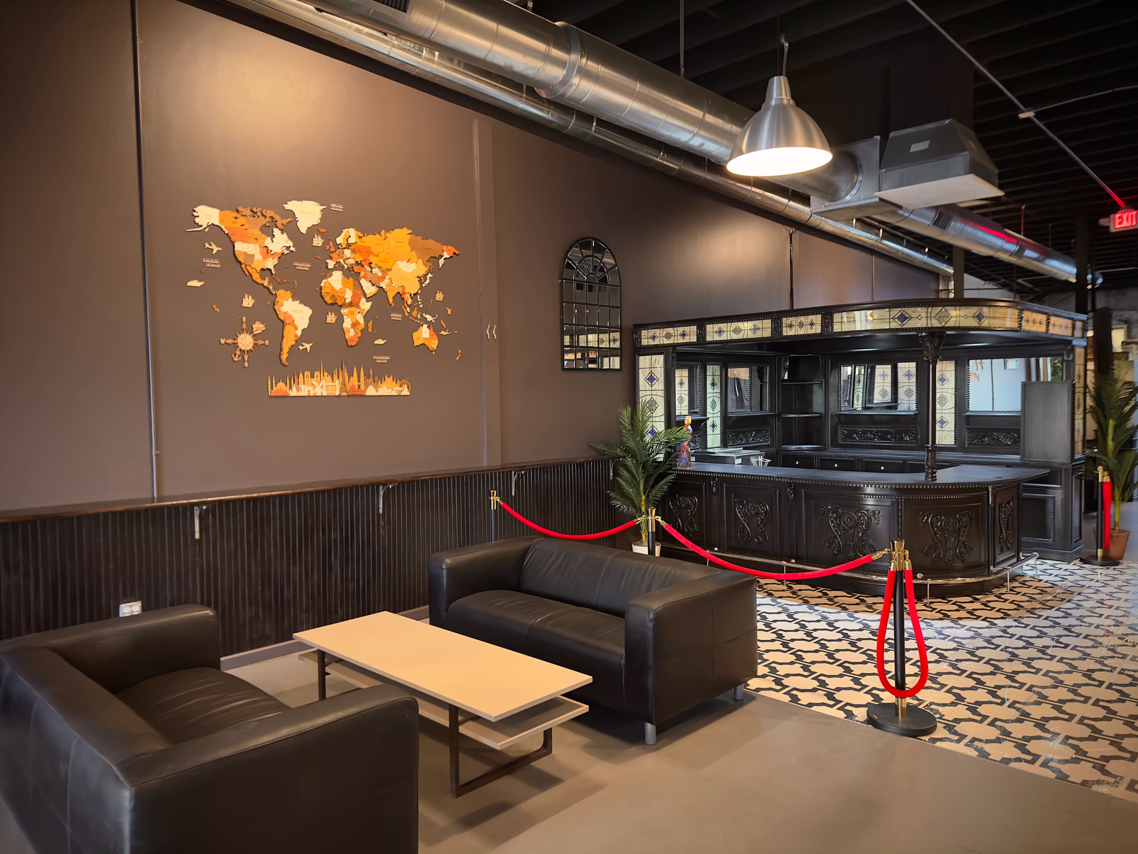A modern interior space featuring two black sofas, a coffee table, a wall-mounted world map, and a bar area with a decorative backsplash, all set against dark walls and patterned flooring.