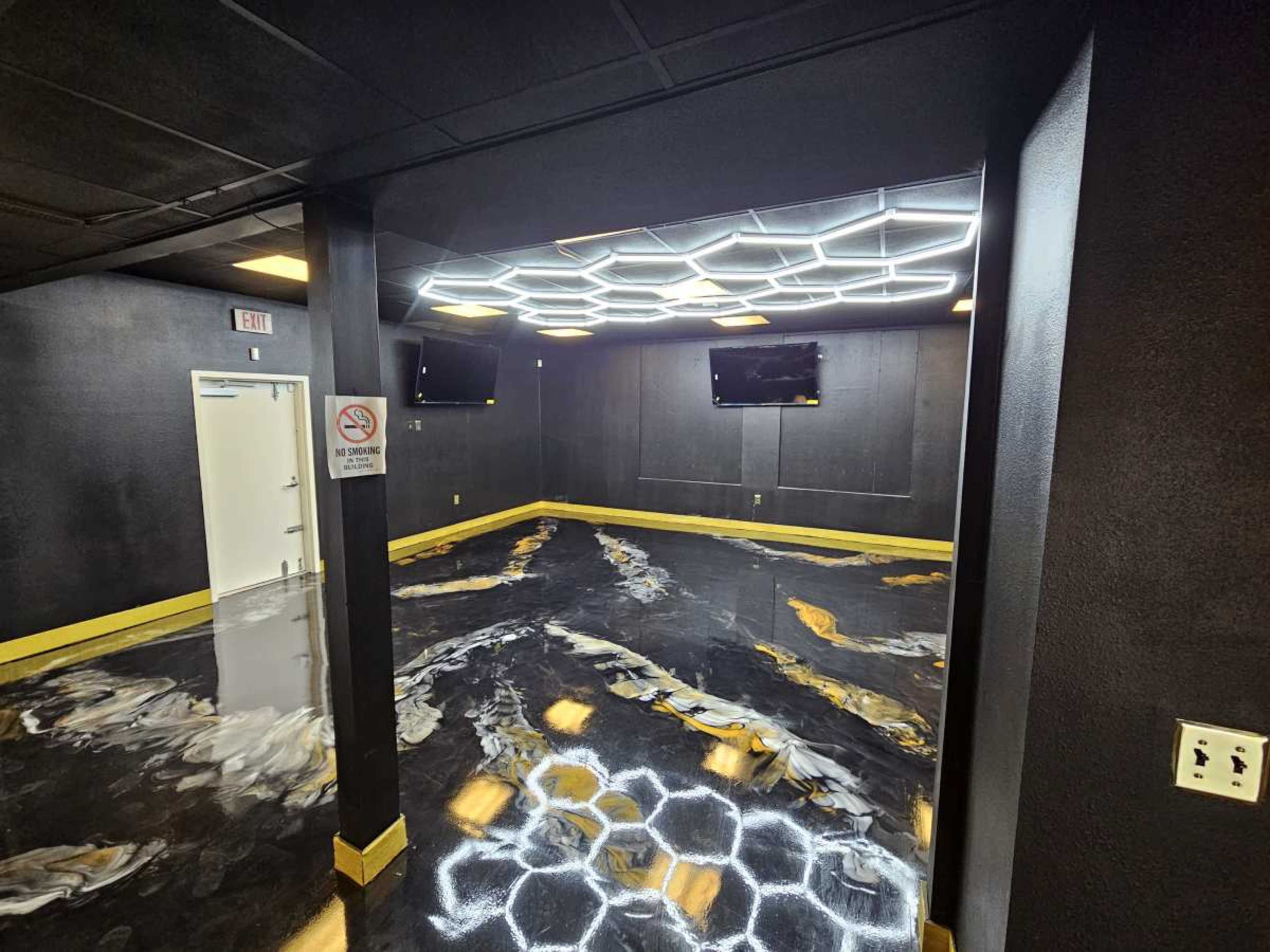 The image shows a room with dark walls, a hexagonal ceiling light design, two mounted TVs, and a glossy black and gold floor.