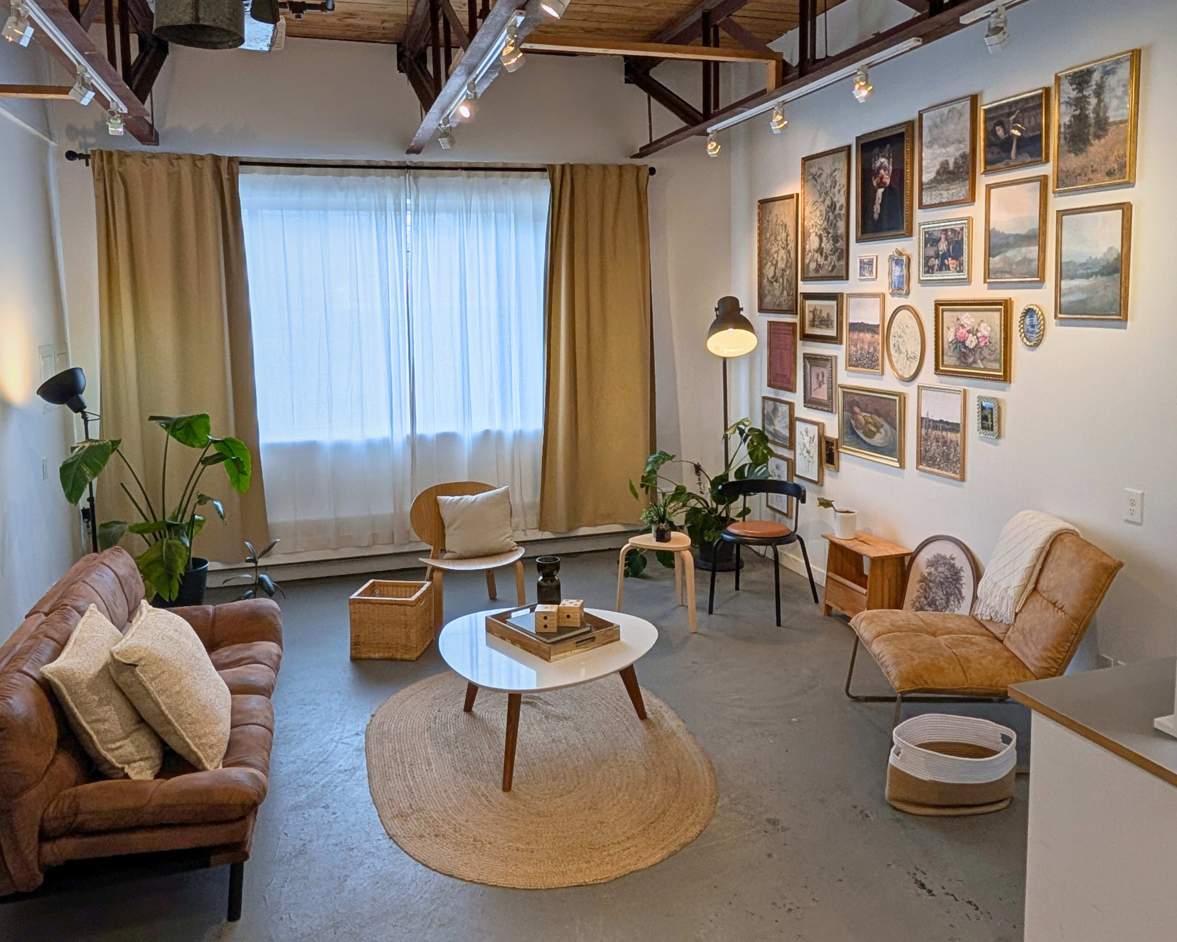 The image shows a cozy living space with a brown leather sofa, a white coffee table, plants, and a gallery wall of framed pictures under soft lighting.