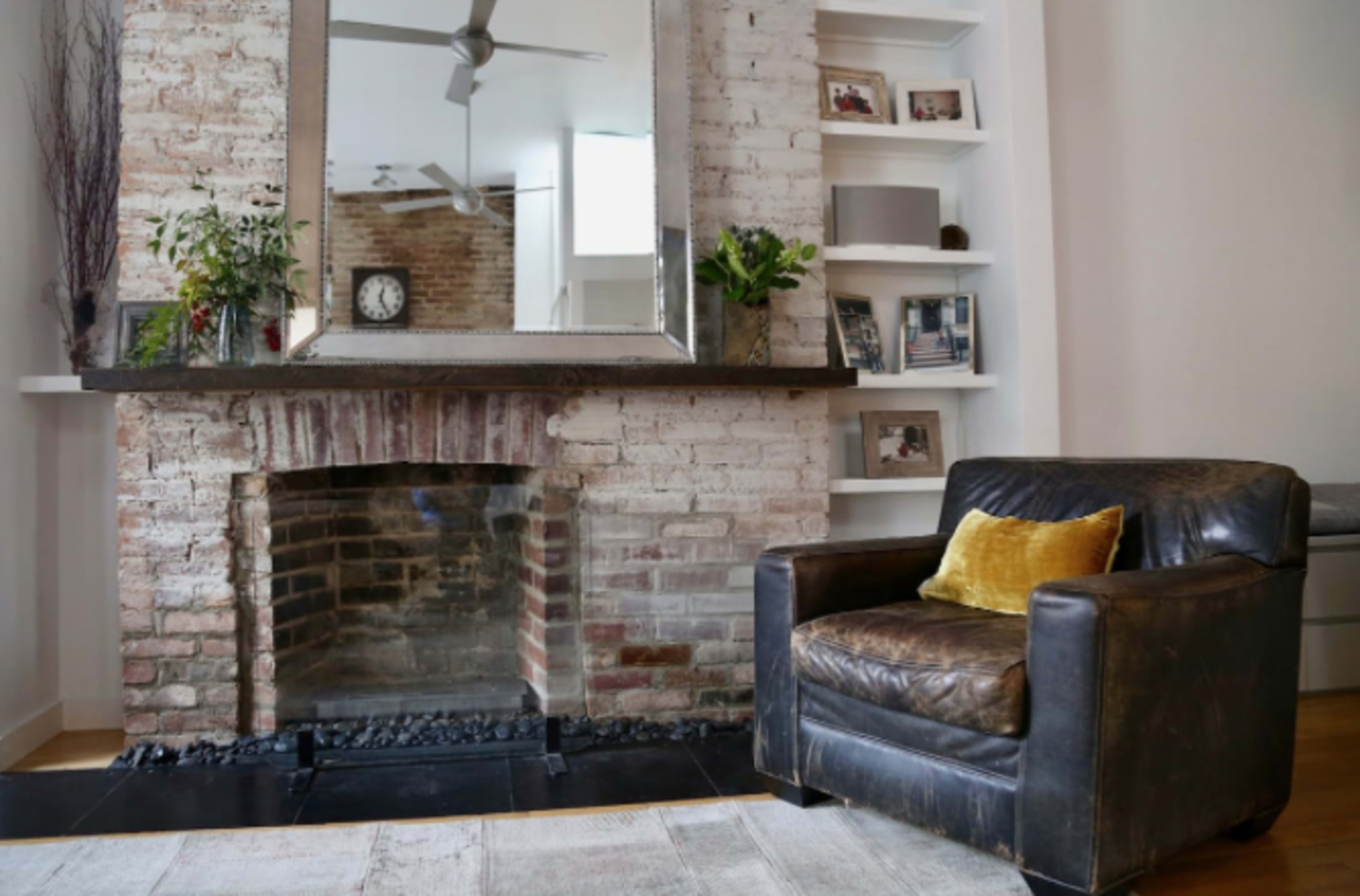A living room with a brick fireplace, a large mirror above the mantel, and a leather armchair adorned with a yellow cushion.