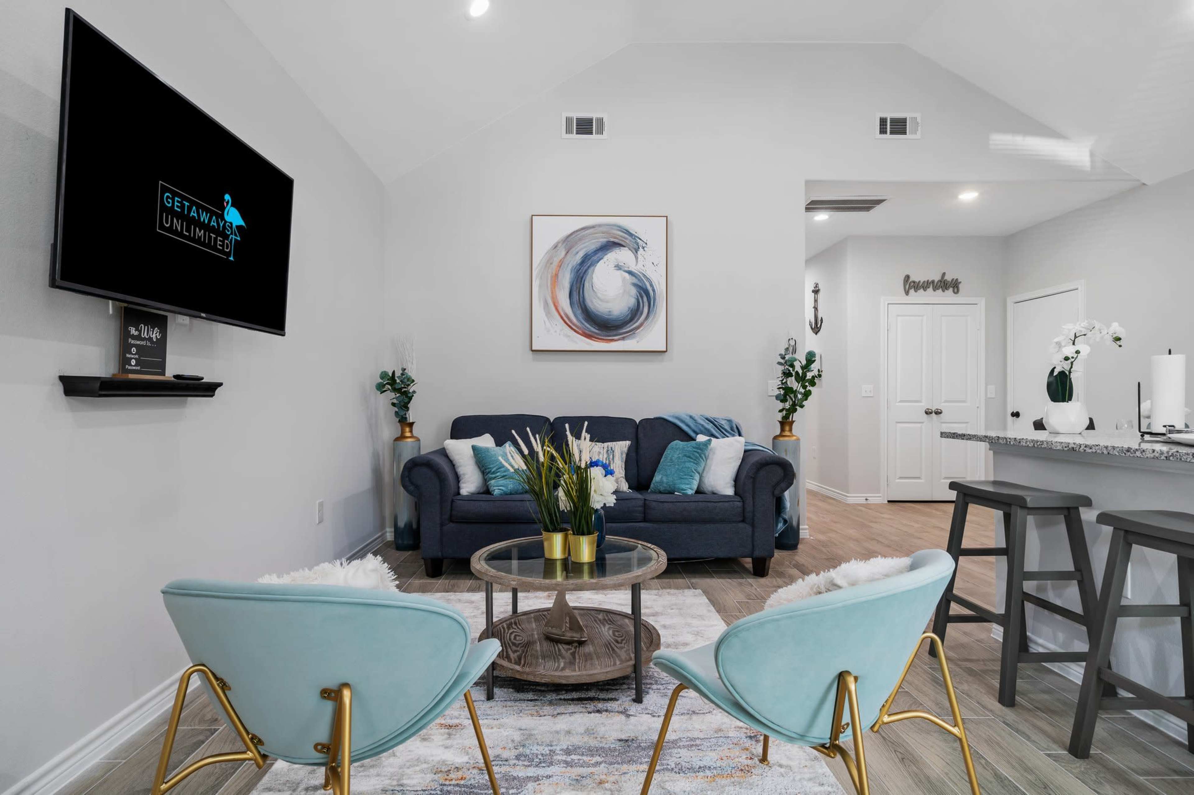 A modern living room features a blue couch and two light blue chairs around a glass coffee table, with a TV mounted on the wall and a painting above it.