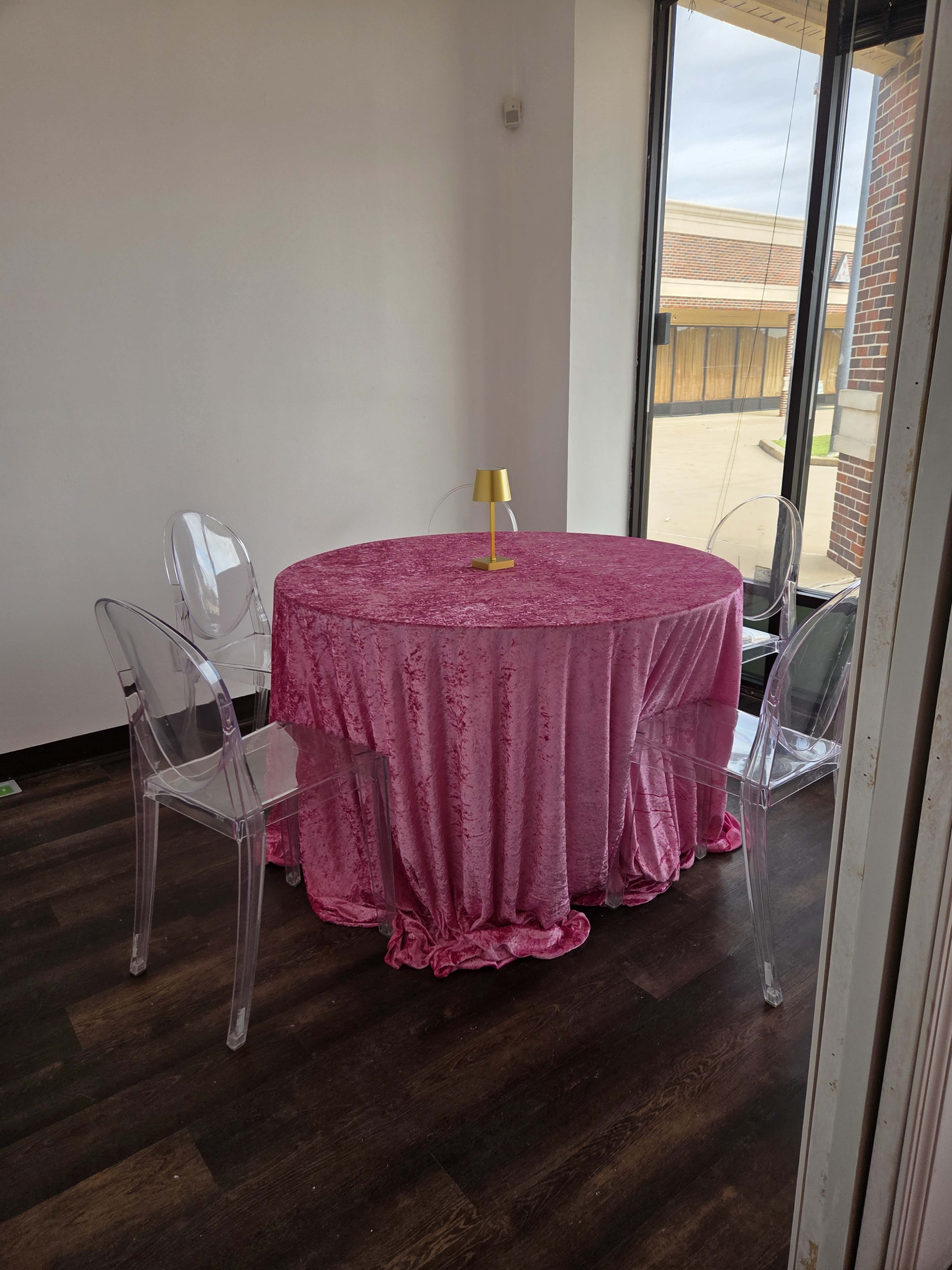 A round table covered with a pink velvet cloth is surrounded by four transparent chairs in a brightly lit room with large windows.