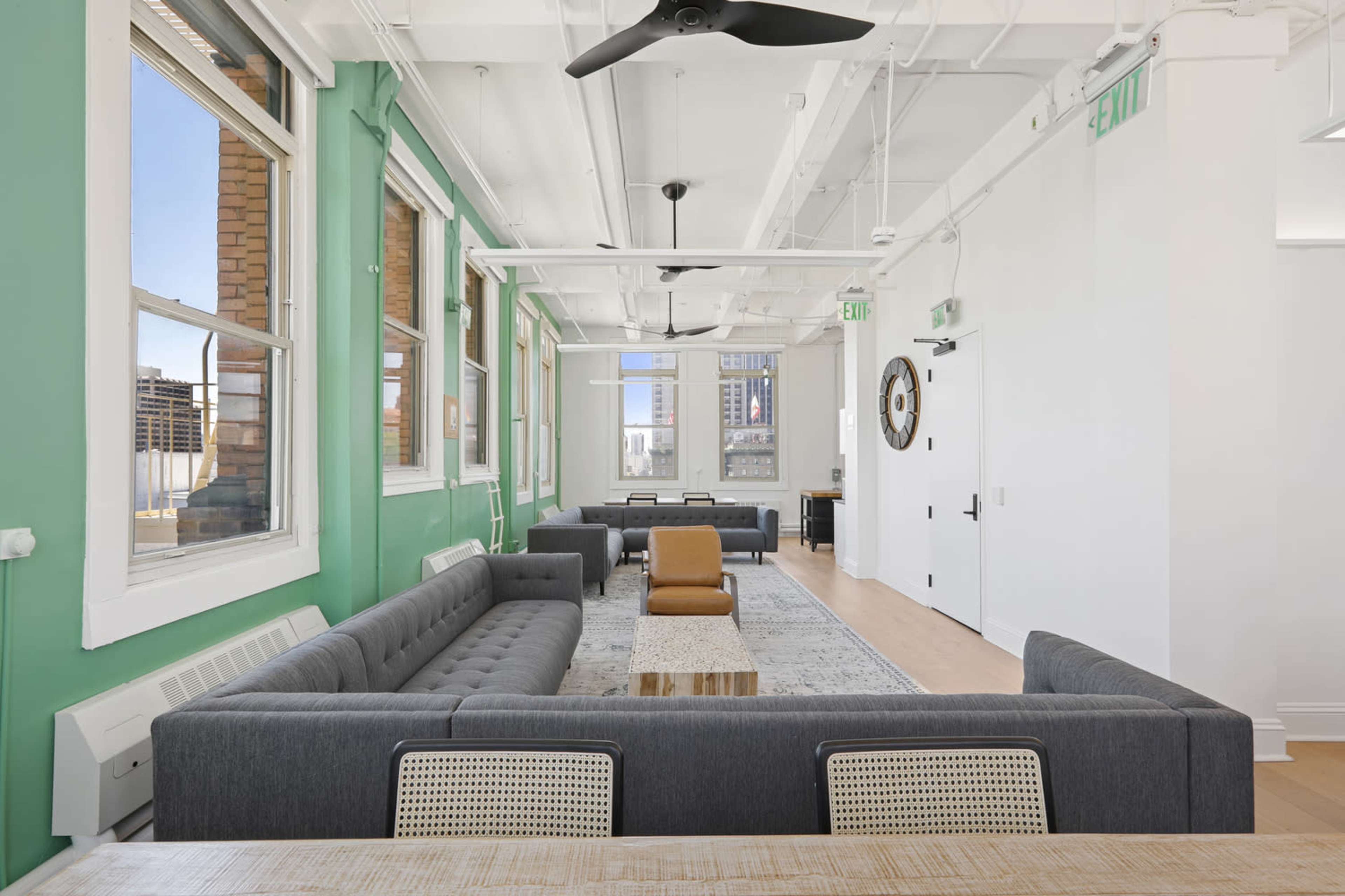 A modern office space featuring a seating area with plush sofas, a coffee table, and large windows allowing natural light to enter.