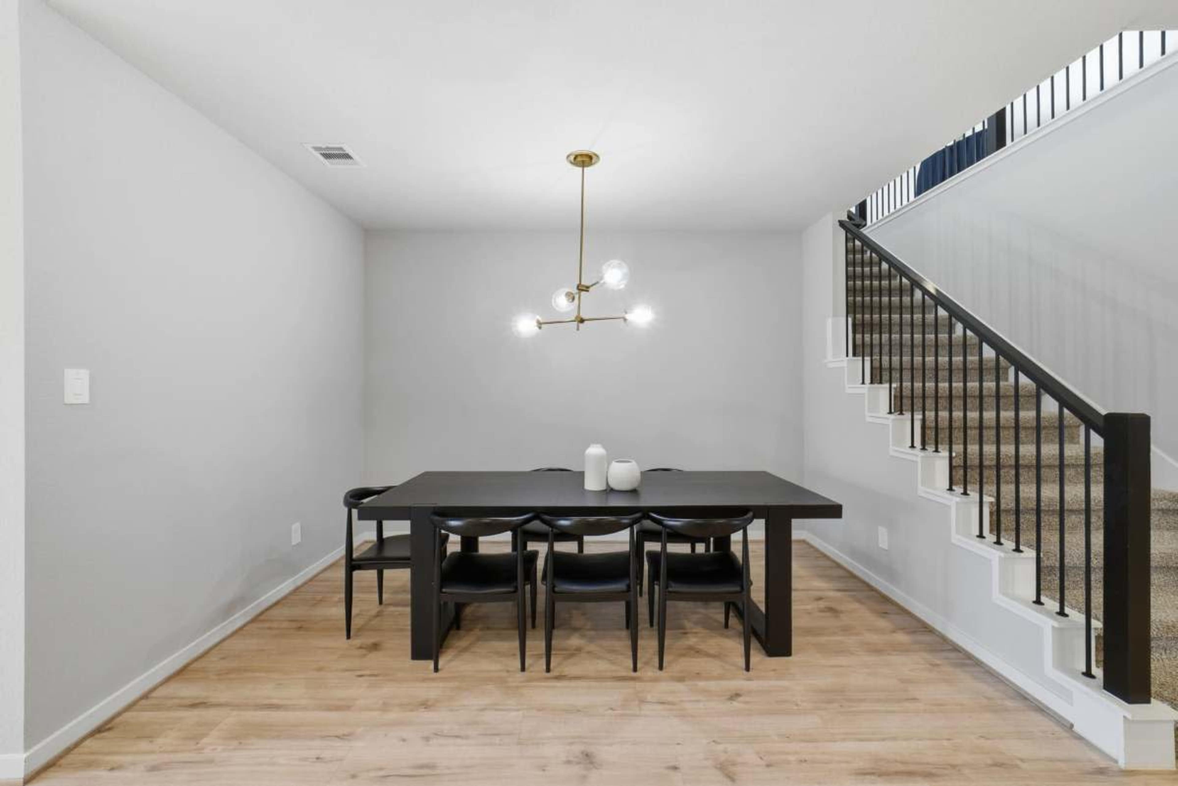 The image shows a modern dining area with a rectangular table and black chairs underneath a light fixture, alongside a staircase with a black railing.
