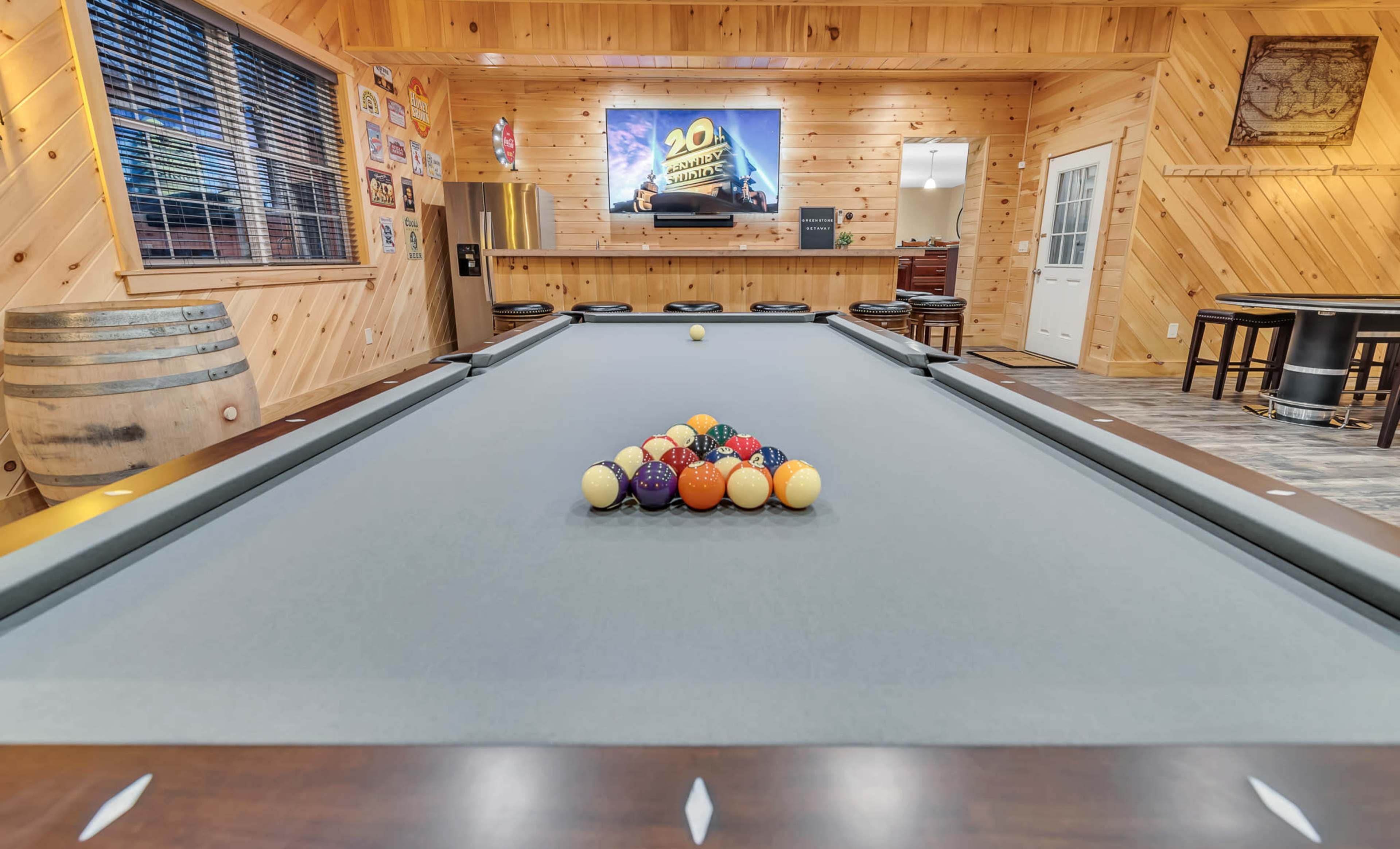 A billiards table with a tight triangle of colored balls is set in a wooden-paneled game room that features a television mounted on the wall.