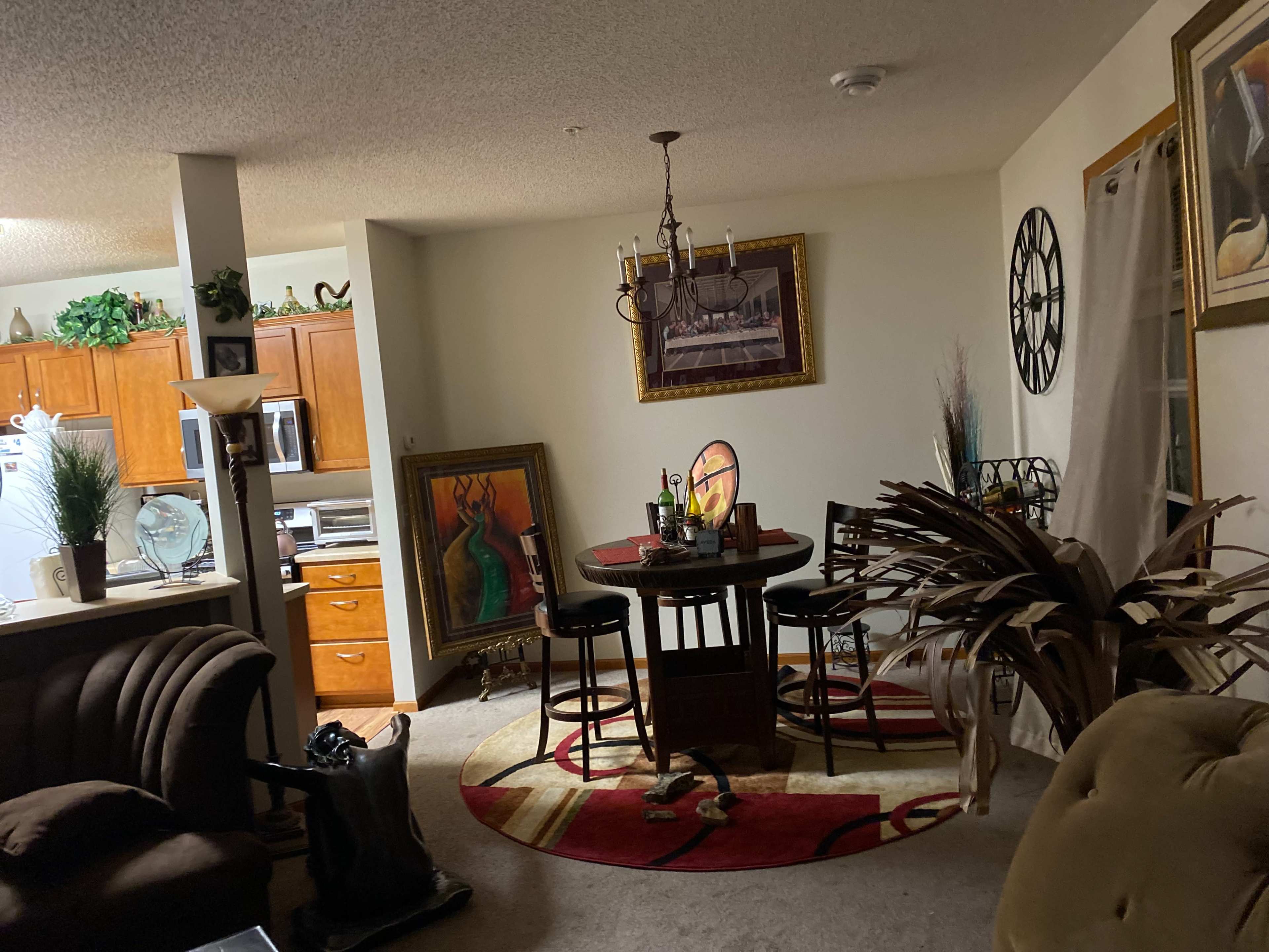 A living room and dining area with a round table and chairs, a large painting, and kitchen cabinets in the background.