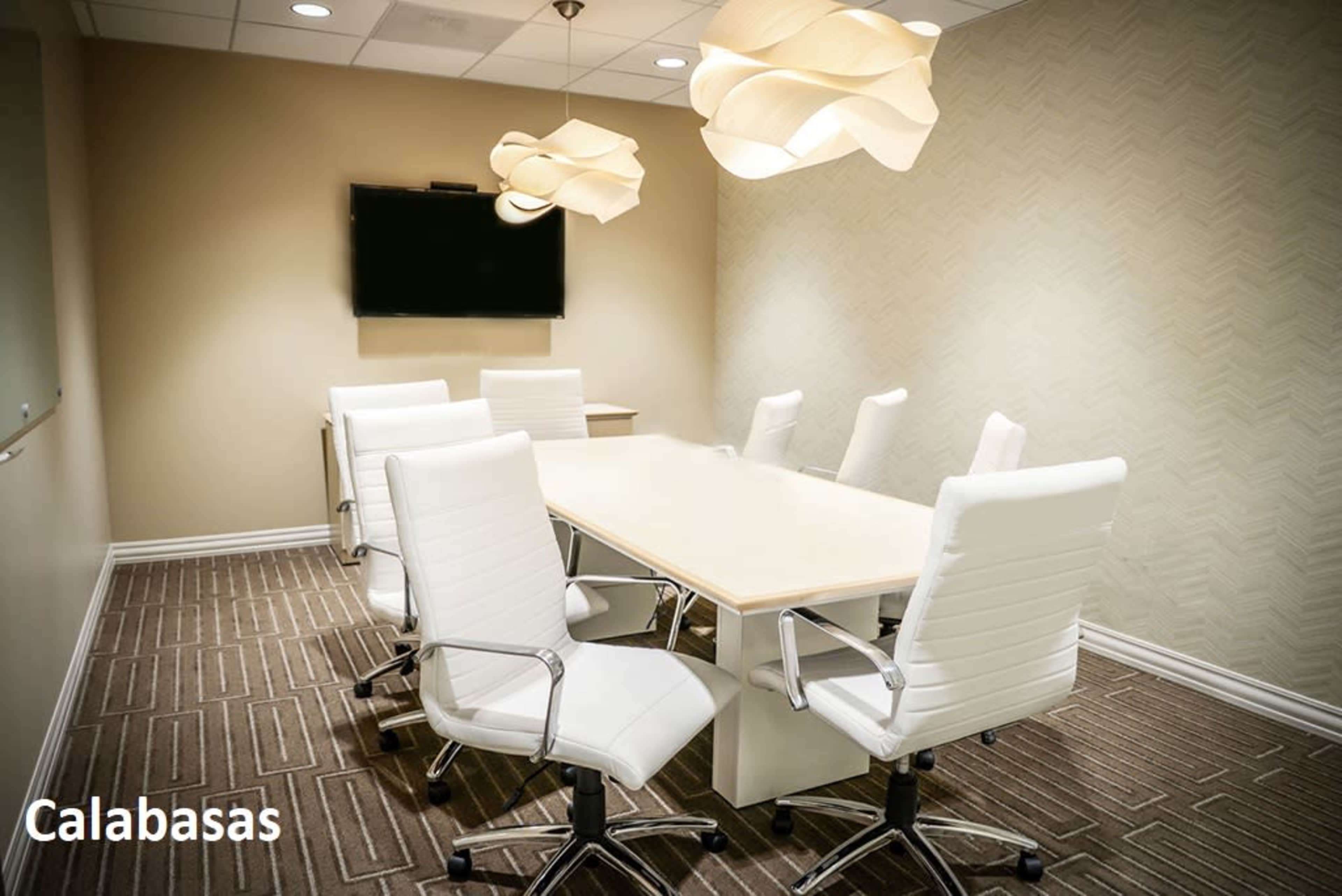 A bright conference room featuring a large table surrounded by six white chairs and a wall-mounted television.