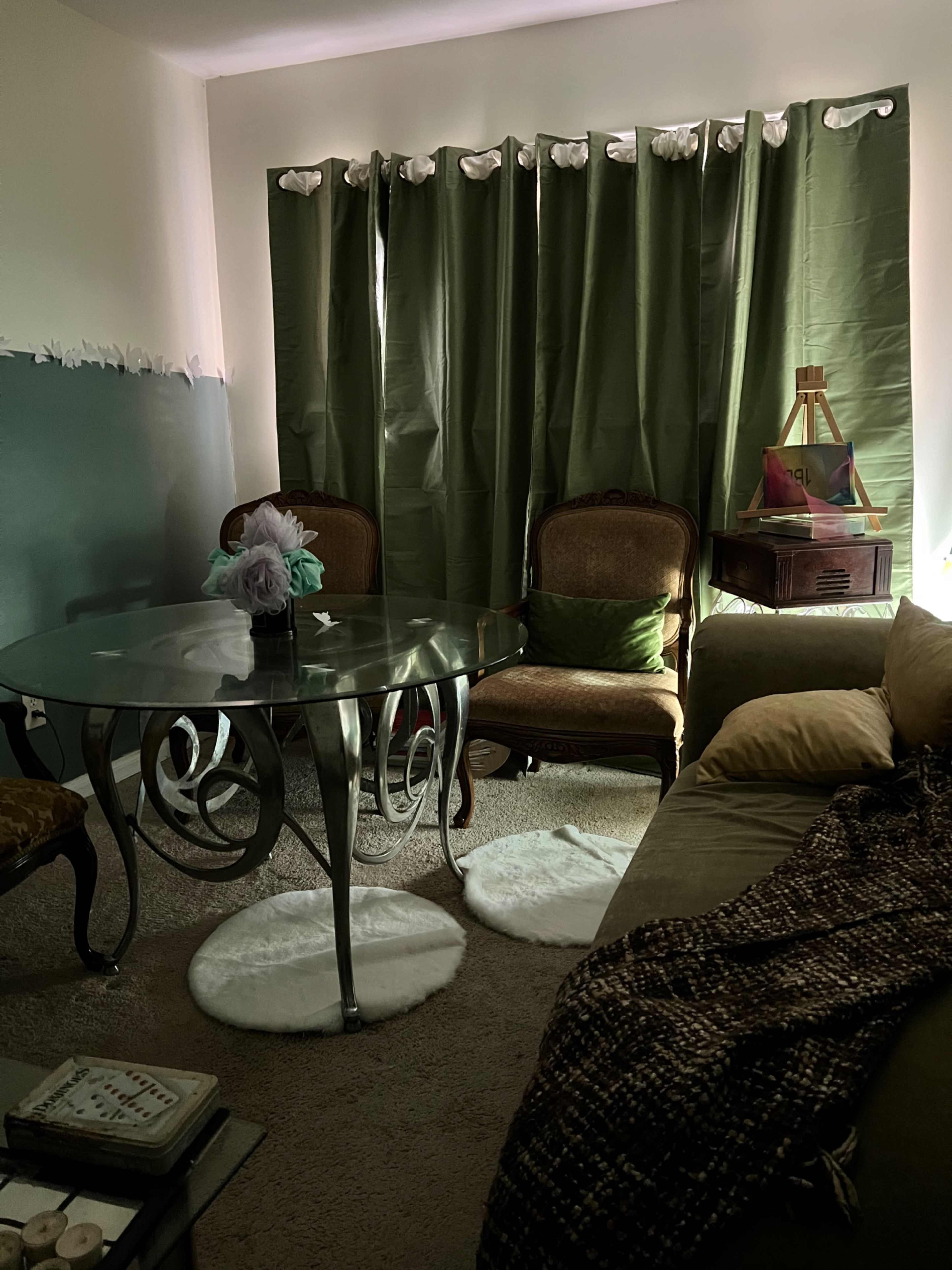 A living room features a glass dining table with curved legs, two upholstered chairs, a couch, and green curtains that block sunlight.