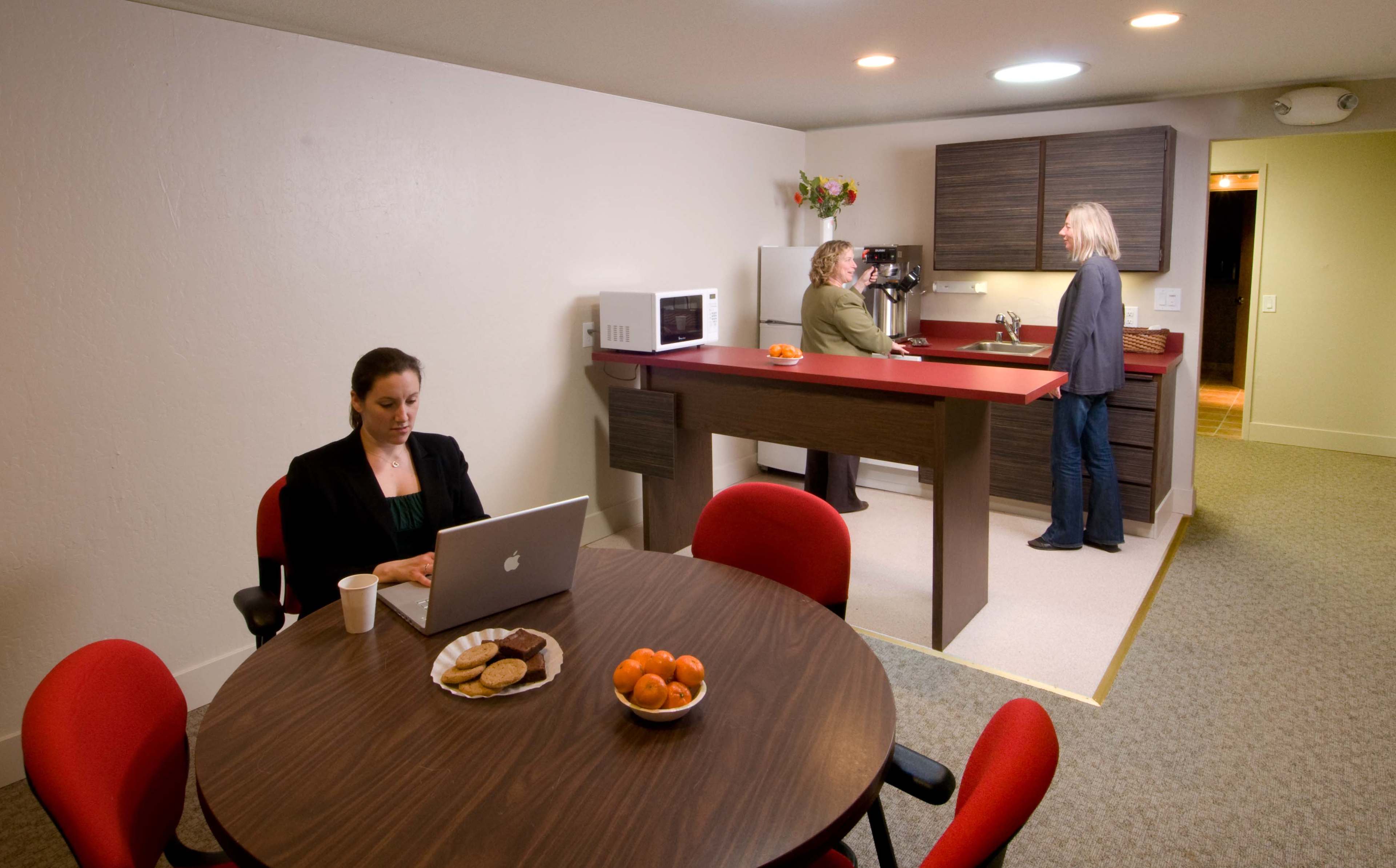 A woman sits at a table with a laptop and snacks, while two other women converse near a kitchen area.
