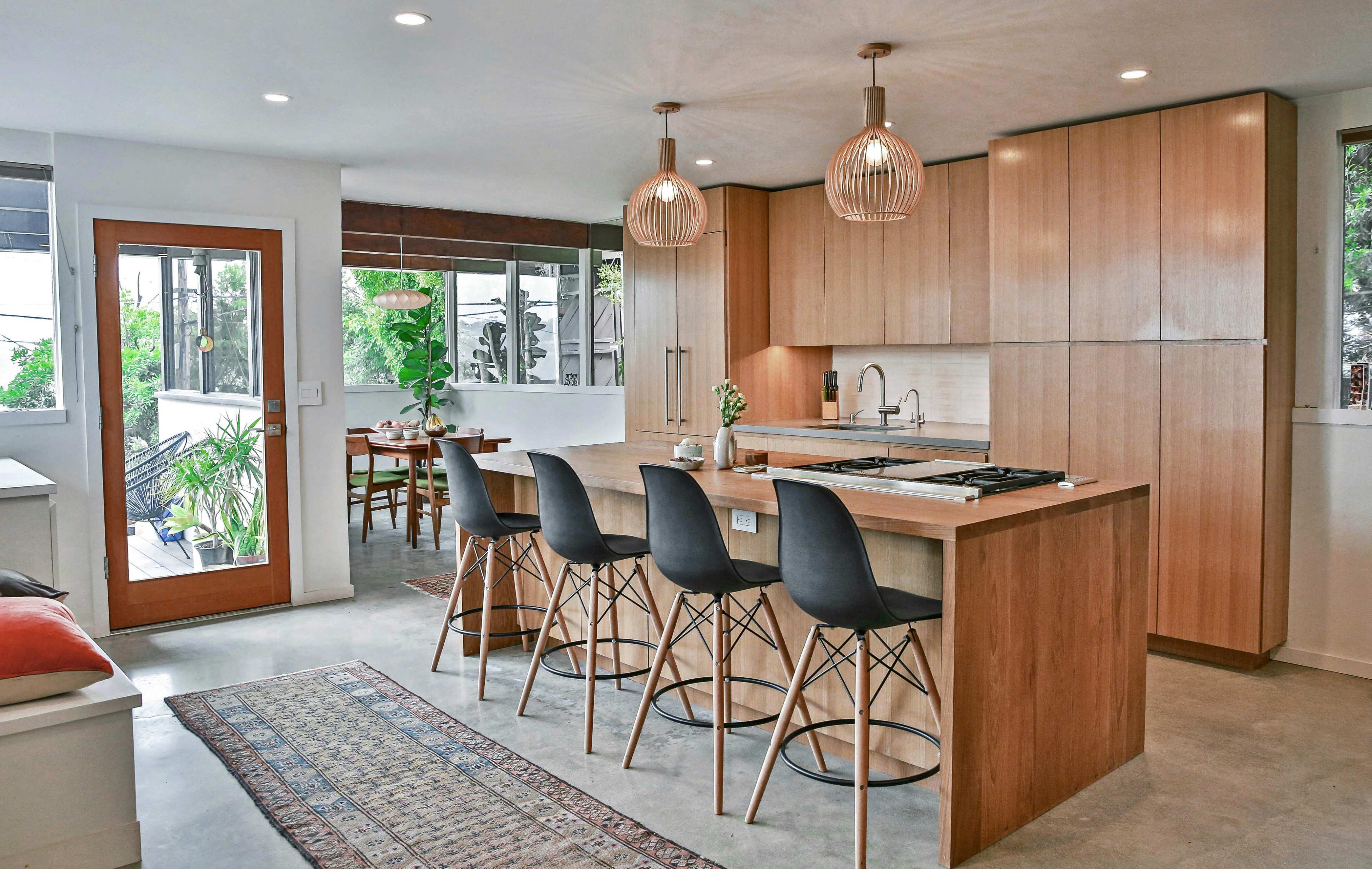 A modern kitchen features a wooden island with seating, pendant lights, and large windows overlooking a dining area.