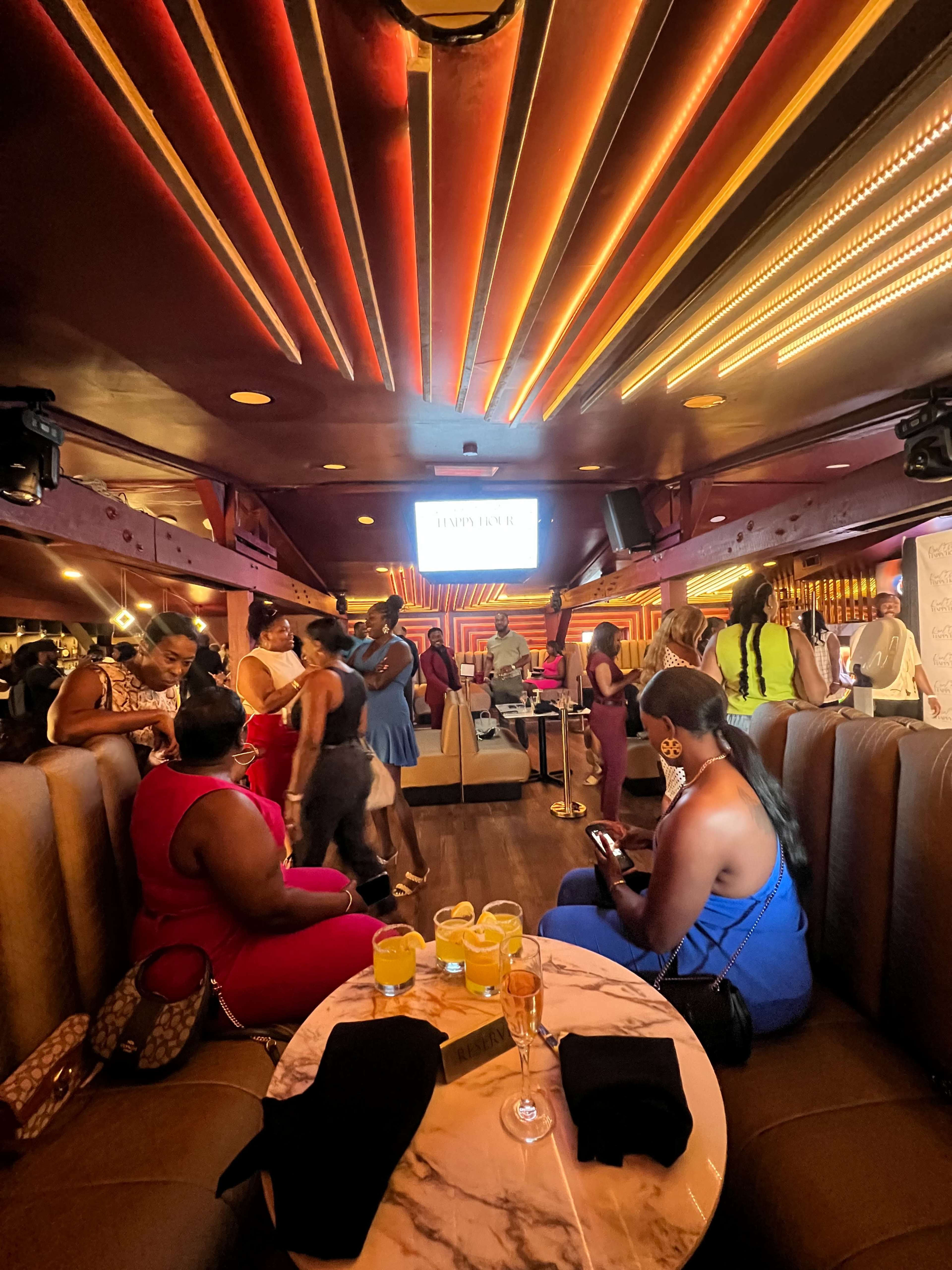 The image shows a lively bar setting with patrons mingling, seated on plush couches, while a screen displays "HAPPY HOUR" in the background.